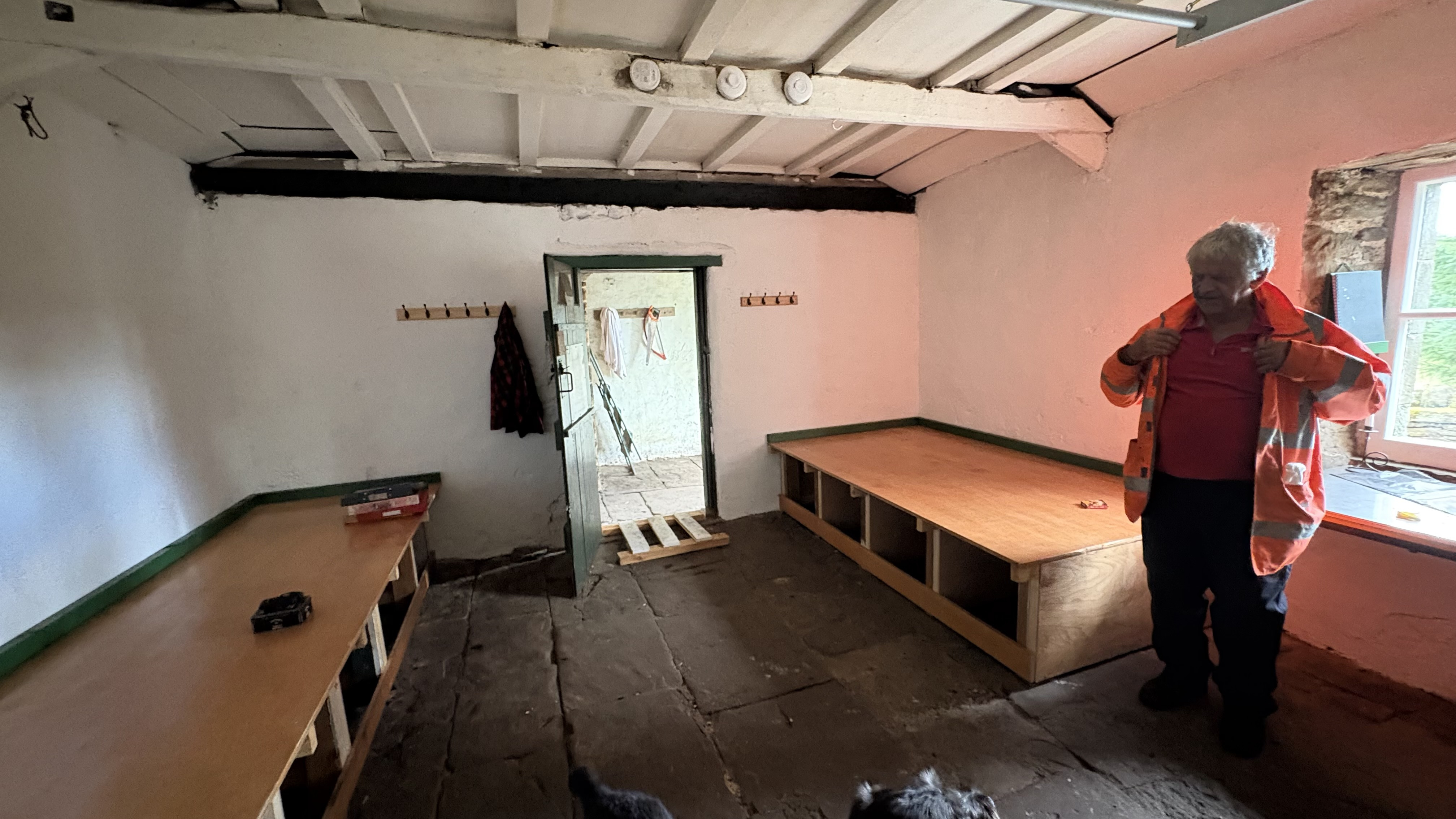 A man in a high-vis jacket standing in the main room of a bothy, white walls, with two sleeping platforms flanking an open door, with a flagged whinstone floor and exposed white-painted beams above.