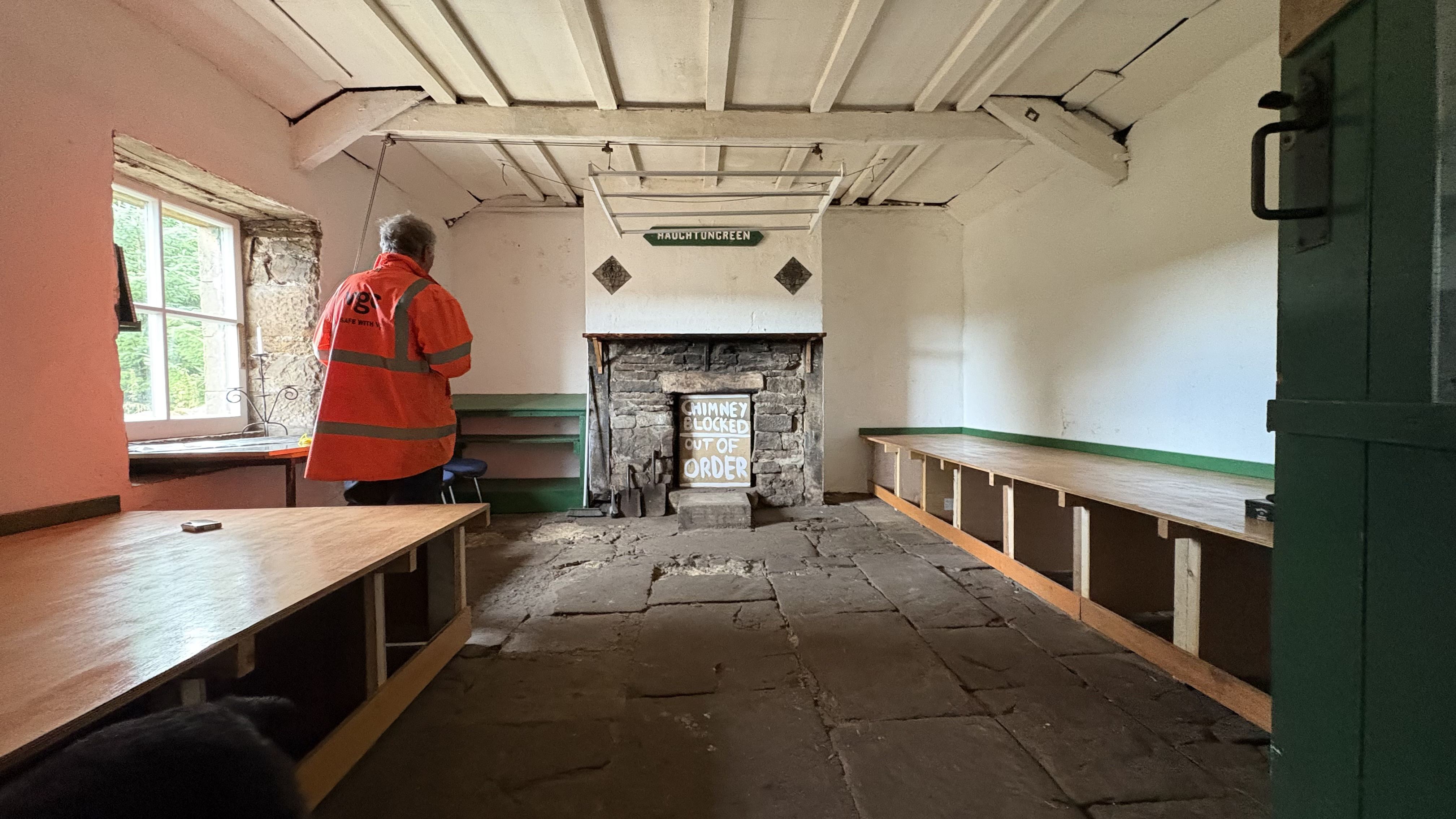 A view down the main bothy room, with flagstone floors and two wooden sleeping platforms against each wall. A stone fireplace at the end of the room is blocked up with an "out of order" sign and a man in a high-vis jacket stands by a window.