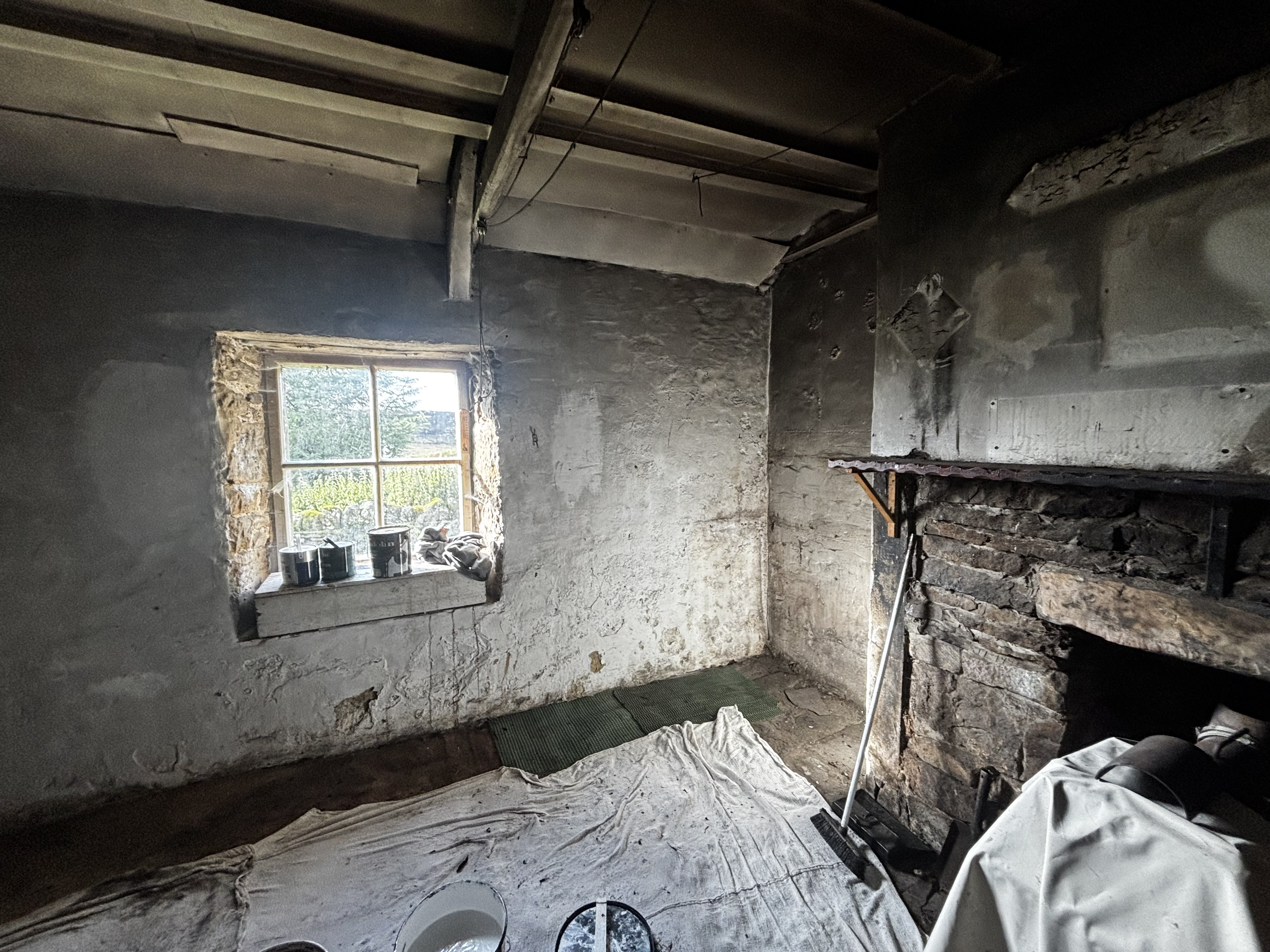 The soot-stained walls and ceiling of the main room of the bothy, with a window looking out on a garden of nettles and a grimy box on the windowsill; a stove is covered by a dustsheet at right.