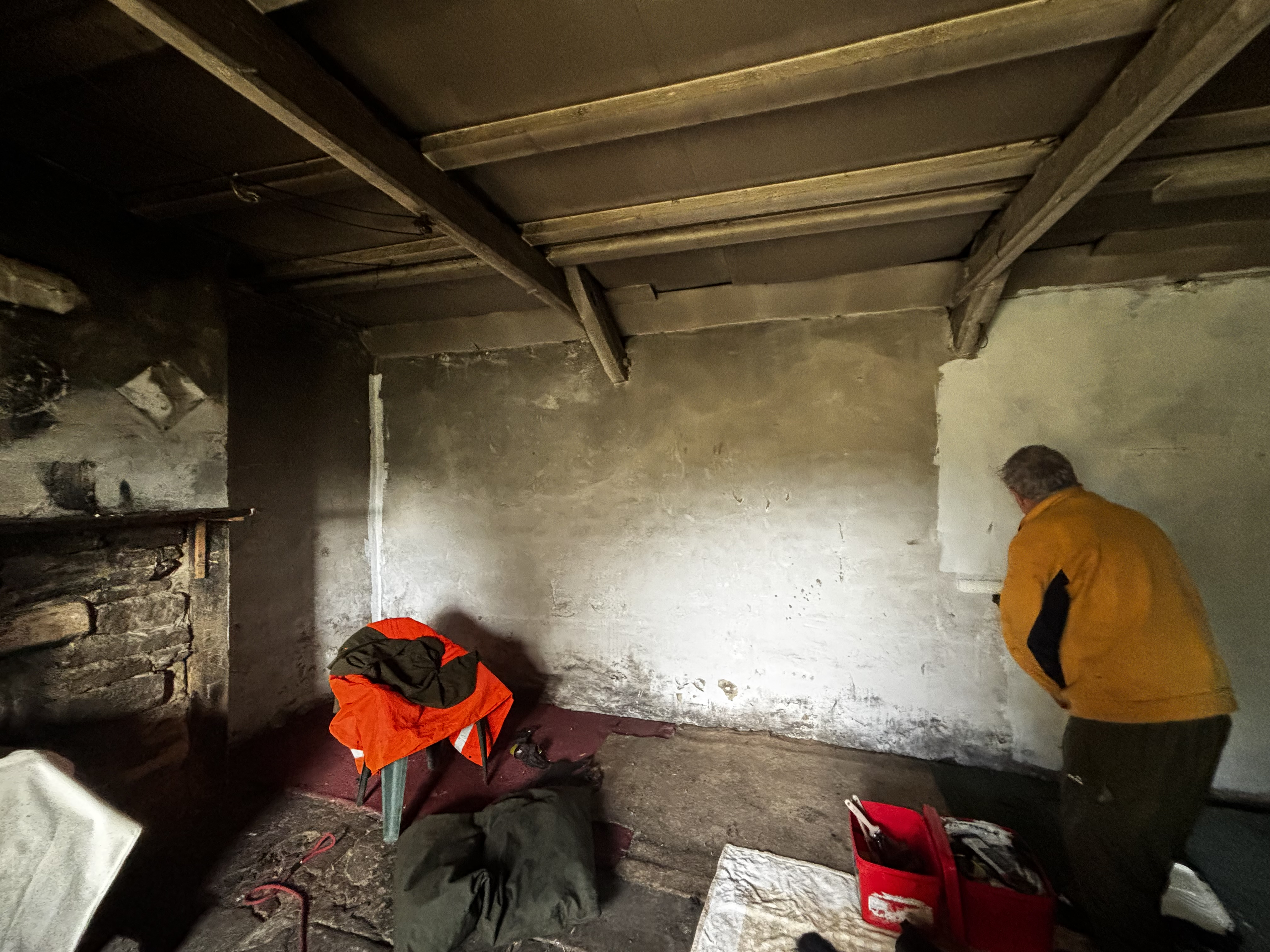 A man in a yellow sweater paints a sootstained bothy wall; dust sheets and carpets are laid on the floor to catch drips; a bare stone fireplace at left is partly covered by a dustsheet. The whole place looks pretty grimy.
