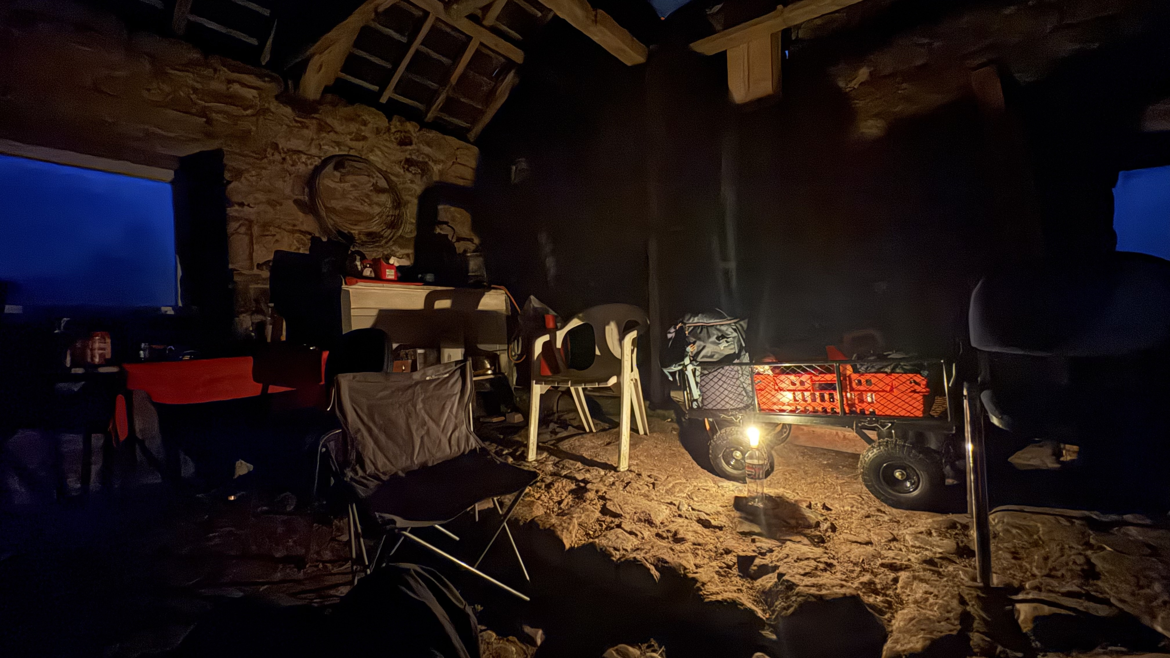 A candlelit view of the byre at the bothy, with a couple of tables up against one wall, a plastic chair and a metal trolley centre, and dark wooden roof beams above.
