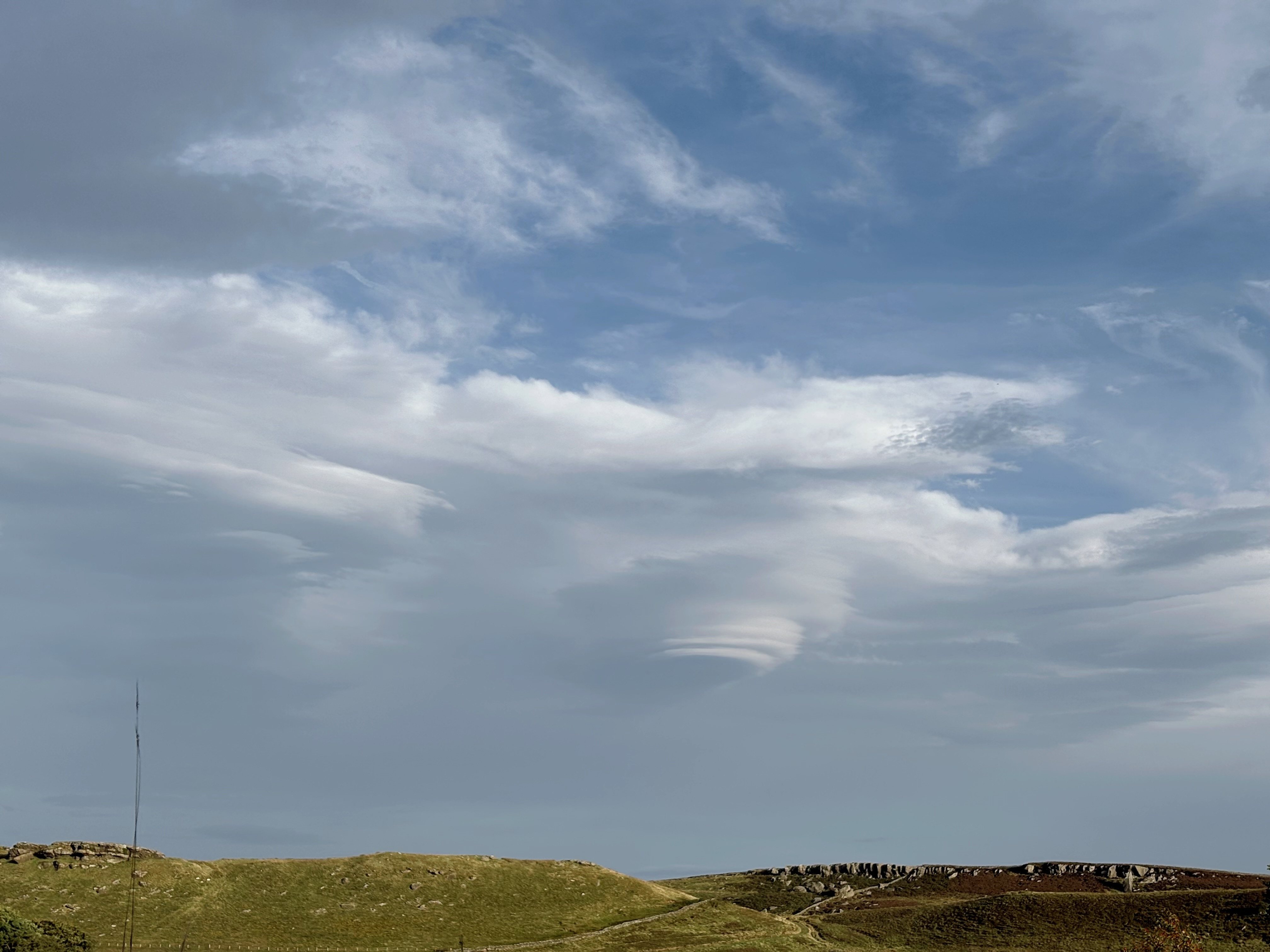 A windswept cloudy sky over a couple of grassy hills strewn with stones; the righthand hill is topped with whinstone crags