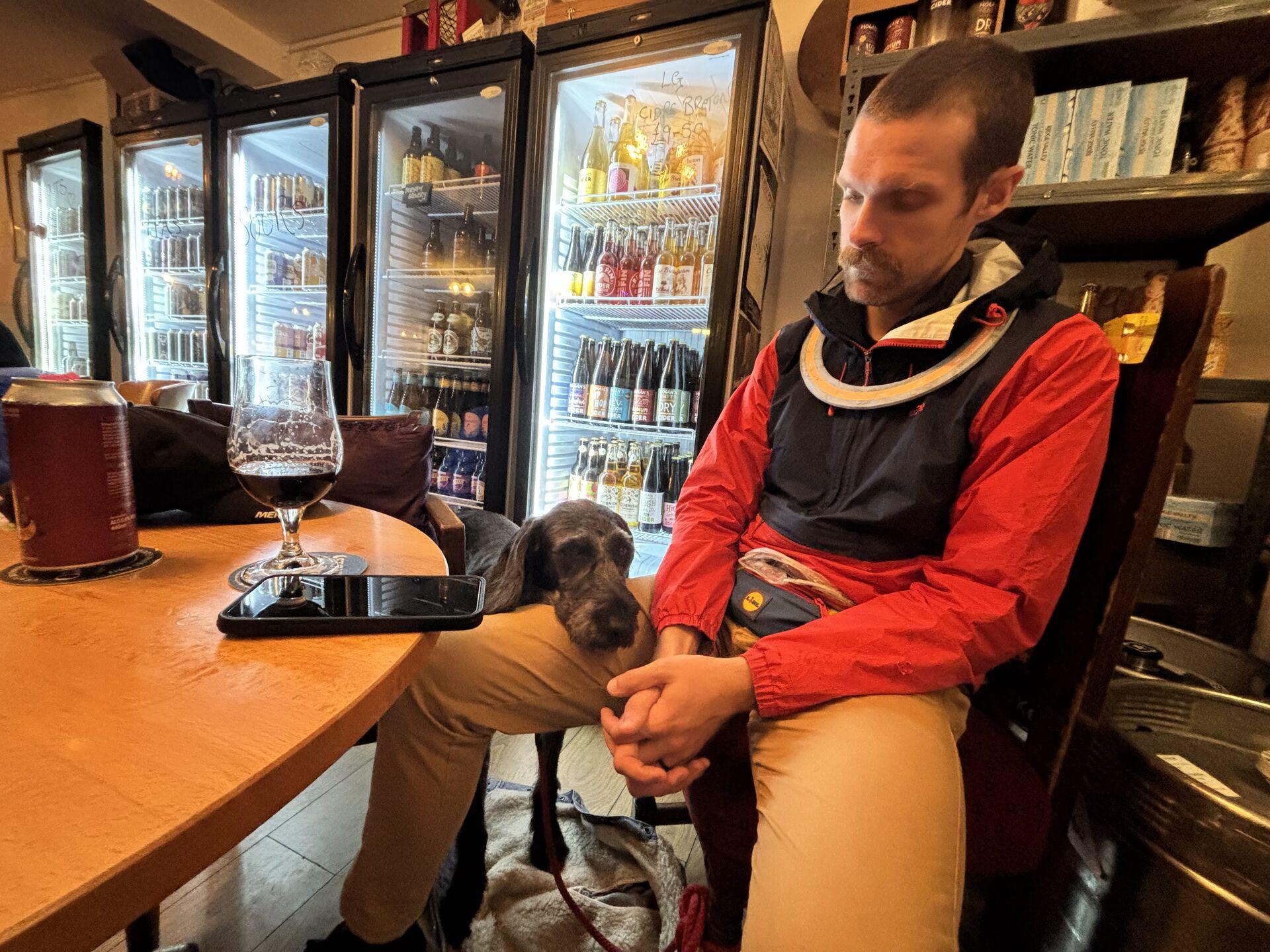 A man in a bar, sitting at a table in front of a row of glass-fronted fridges lined with expensive-looking beers; a dog is resting its head on the man's leg