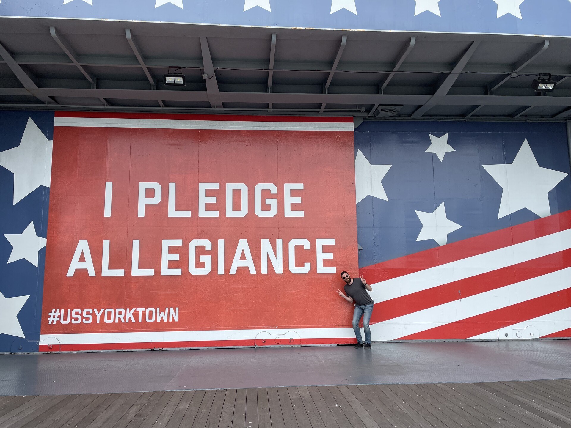 The author making a cutesy double-peace-sign gesture in front of a massive banner reading "I pledge allegiance" with requisite stars & stripes.