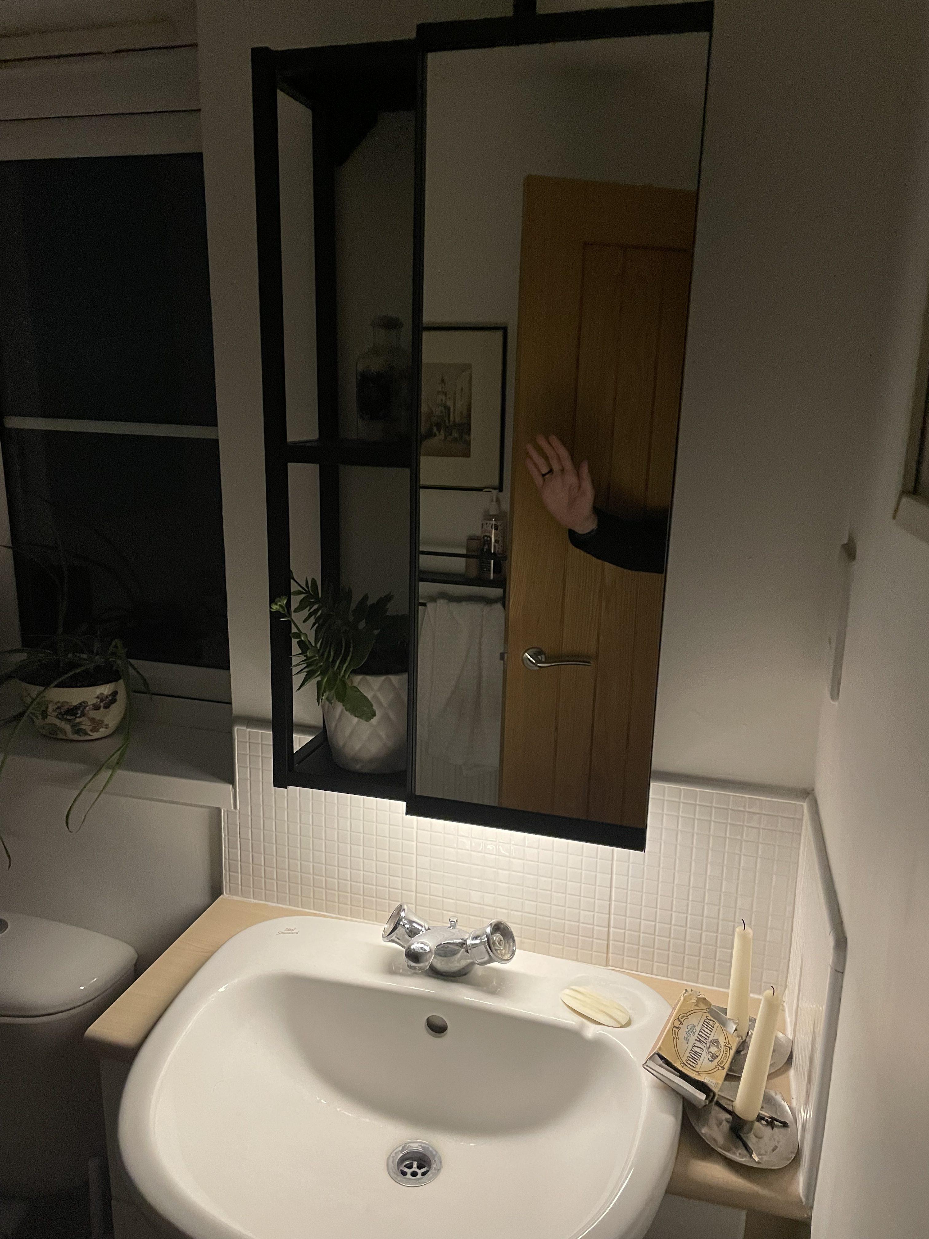 A dimly lit bathroom scene featuring a white sink with a chrome faucet, a soap dish, and two candles on the counter. In the background, there is a mirror with a wooden door partially visible and a hand raised in a wave. A plant is situated on the edge of the sink, and a framed picture hangs on the wall beside a towel rack. A window can be seen with dark surroundings outside, adding to the ambiance.