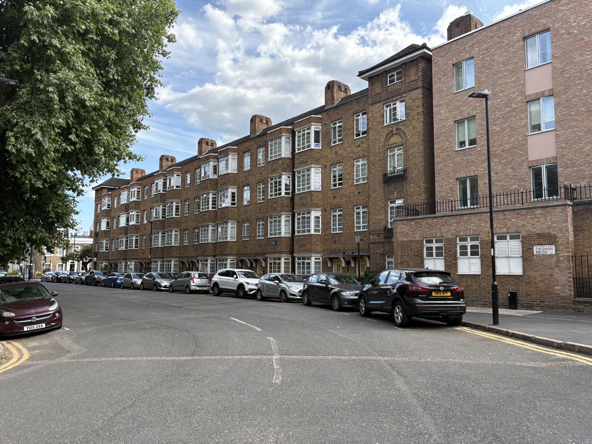 Street view of a row of parked cars along a quiet road lined with multi-story brown brick apartment buildings under a partly cloudy sky. Sign reads "Coleman Fields N1."