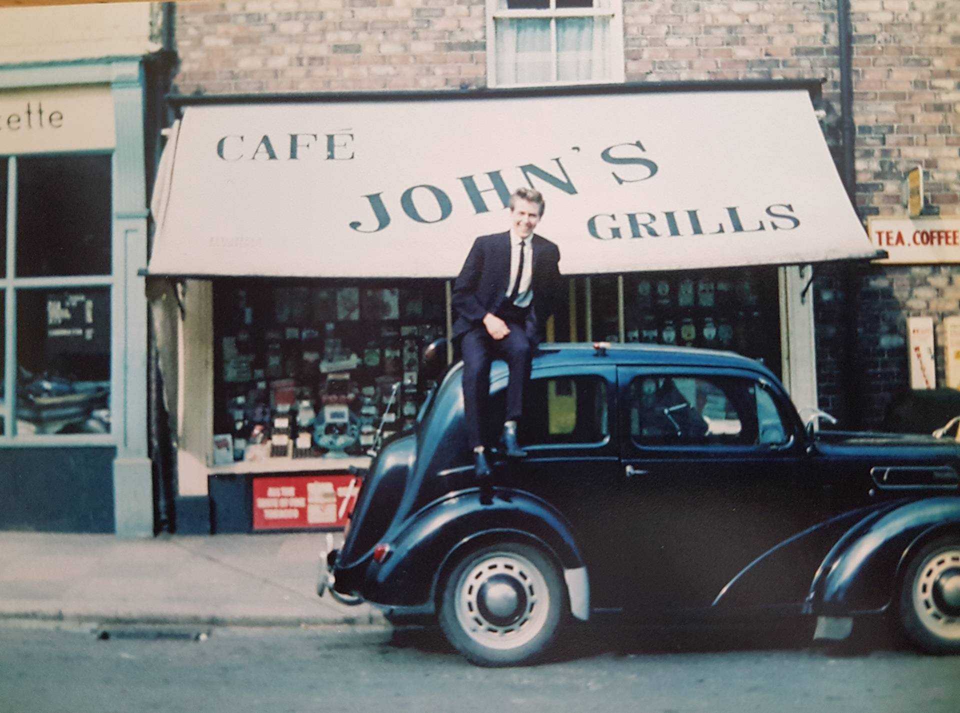 A 1960s photo of John's Cafe, situated on the bottom floor of an old row house, with an old-timey car parked outside and a man in a suit sitting on its roof