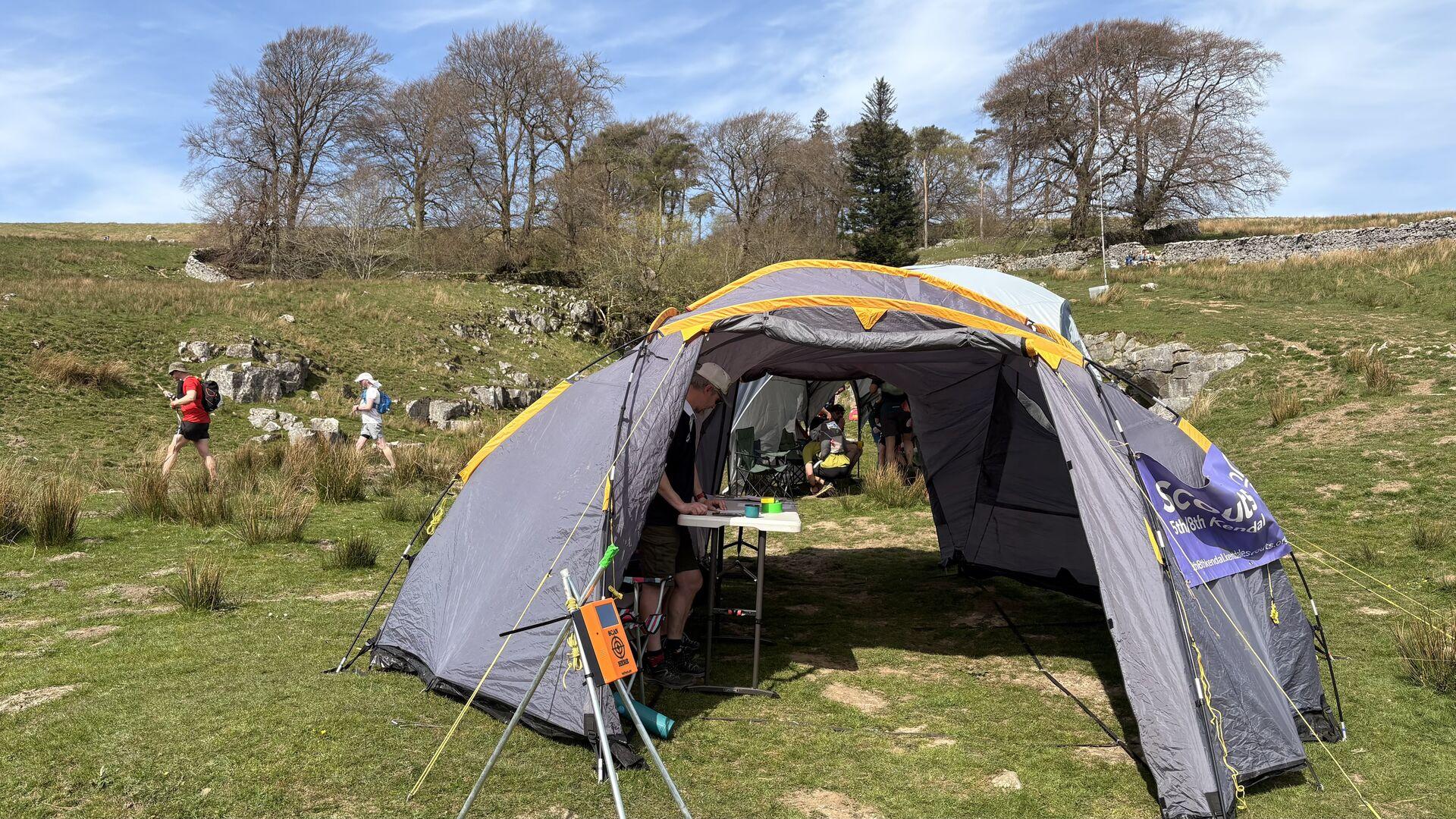 A grey tent pitched on a grassy clearing, with a grassy hill rising behind, with some bare trees enclosed by a stone wall halfway up the slope