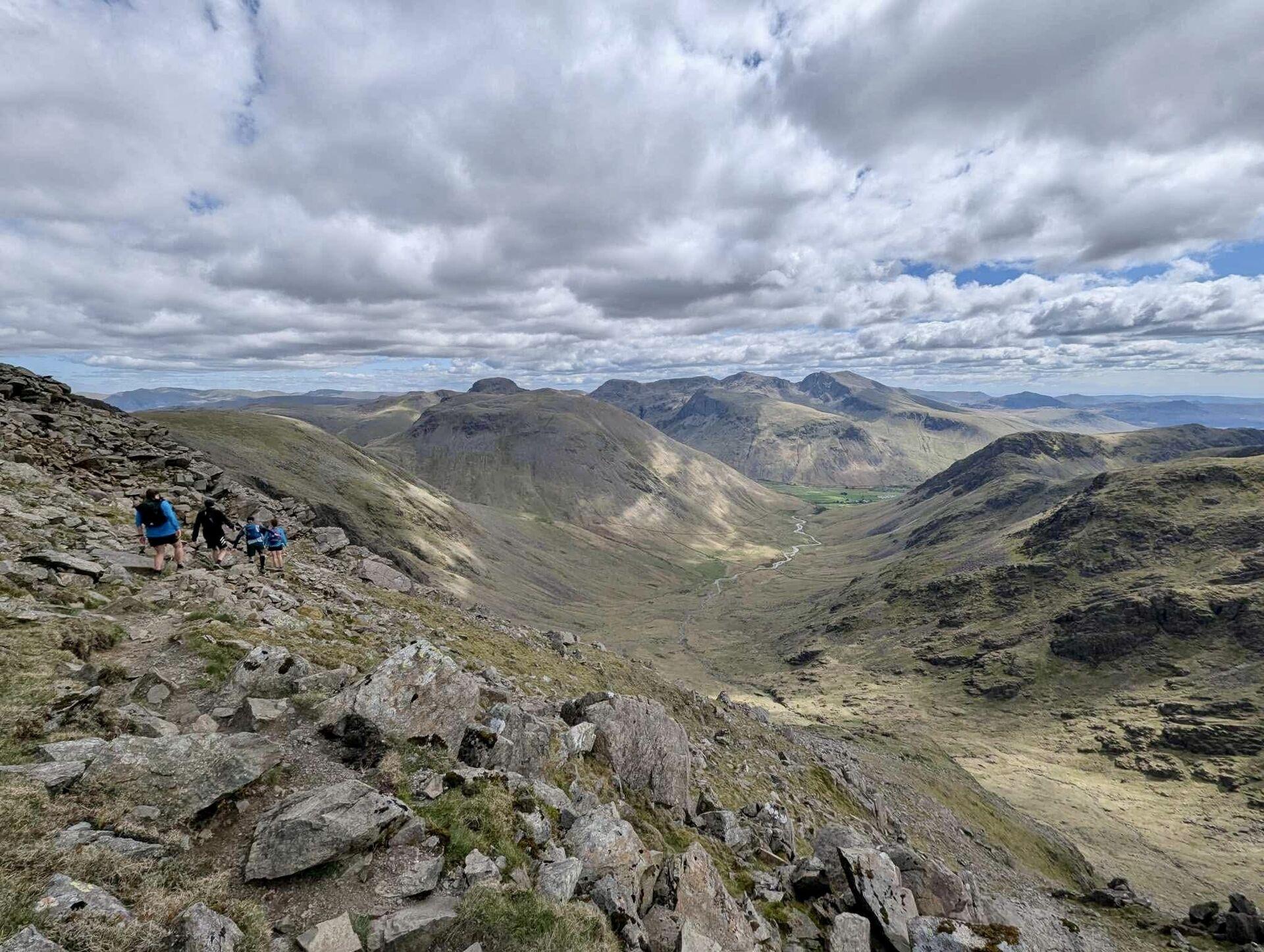 A view down Mosedale, with four runners picking their way through the boulder field on the lefthand slope, looking across to Kirk Fell and Great Gable in the distance
