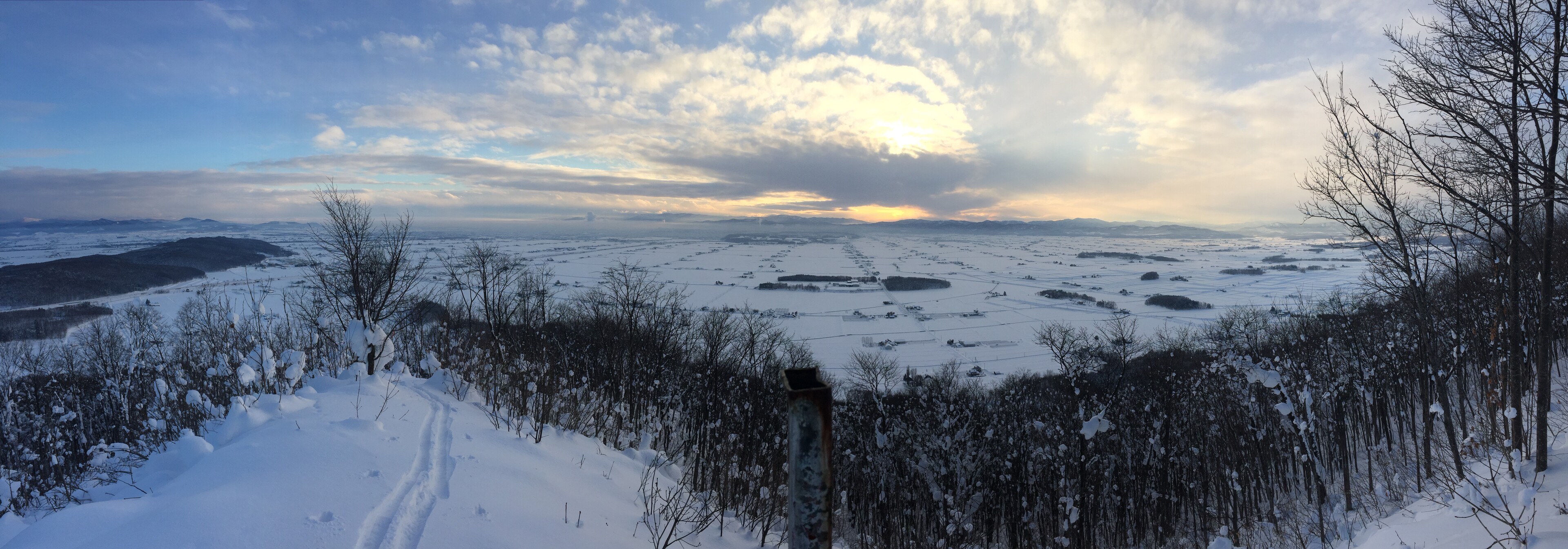 A panorama from the summit of the hill: snow and bare trees below, with my ski tracks coming up from bottom left. A late sun setting behind some clouds in the distance.