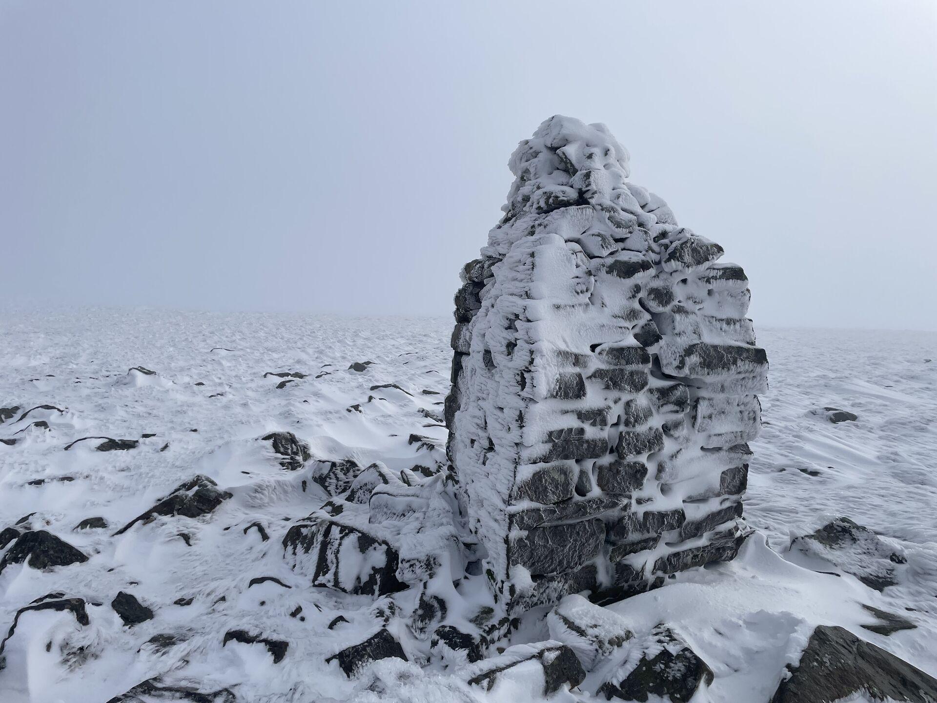 A snow-covered stone cairn, known as Knock Old Man, stands on rocky terrain blanketed in snow. The cairn is partially obscured by frost and ice, and the background features a misty, white landscape, suggesting harsh weather conditions.