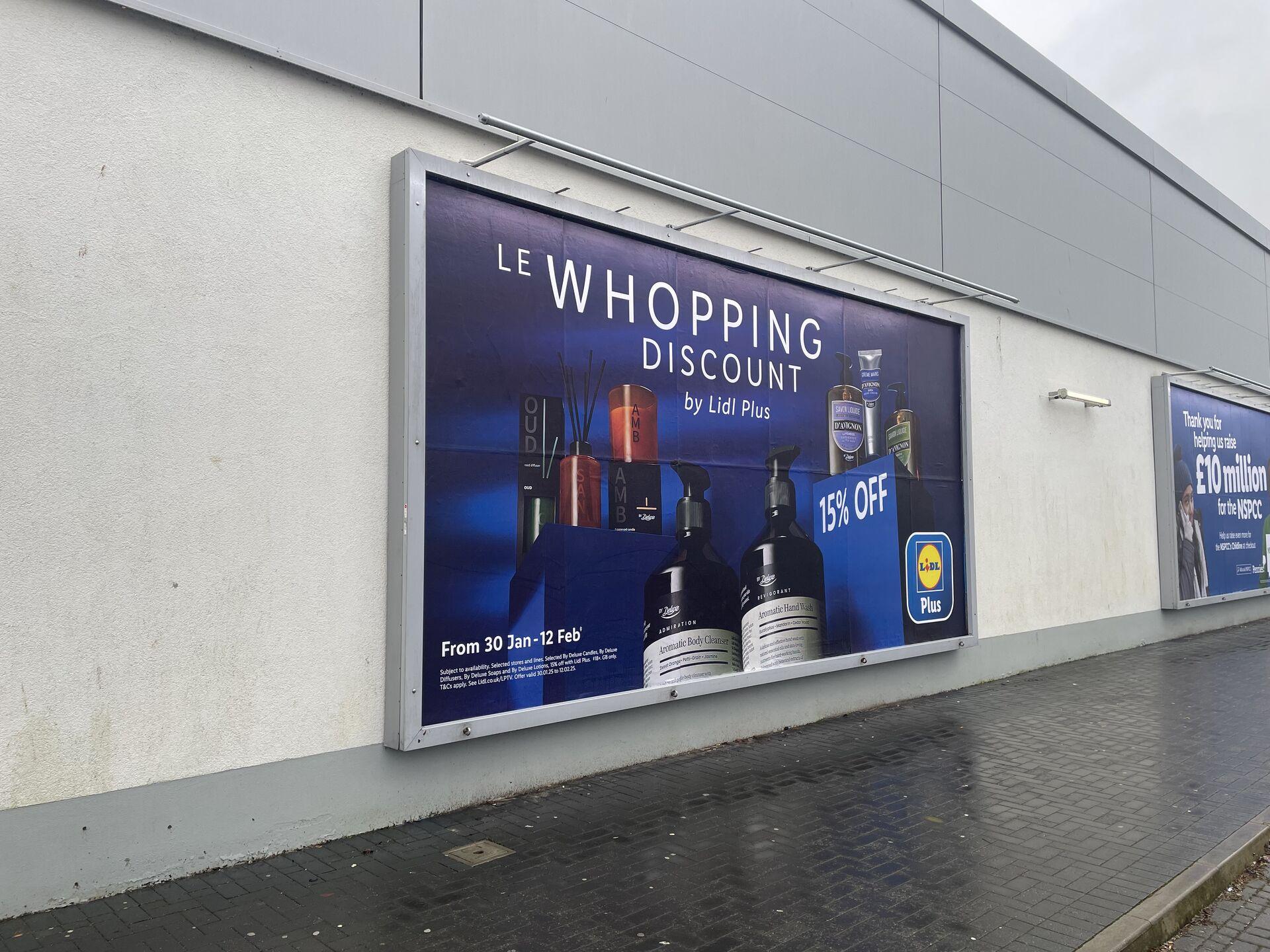 A large advertisement displayed on a wall outside a building. The ad features the text "LE WHOPPING DISCOUNT by Lidl Plus" in bold letters, with "15% OFF" highlighted. Below, it notes the promotional period of "From 30 Jan - 12 Feb." The background is dark blue, and images of various products, including scented candles and lotions, are visible. Another smaller sign on the adjacent wall expresses gratitude for fundraising efforts, mentioning £10 million for the NSPCC. The surrounding area includes a paved walkway and a gray building wall.