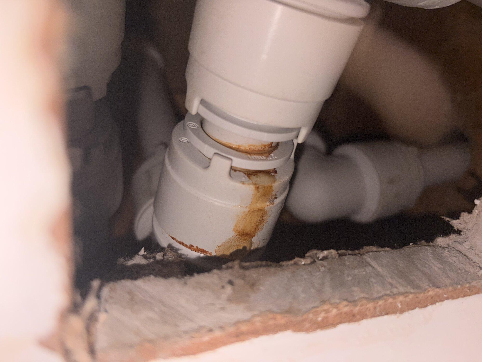 A closeup of a plastic speedfit plumbing fitting, with a stripe of brown rusty water leaking out of one side. Other plastic pipes are out of focus in the background