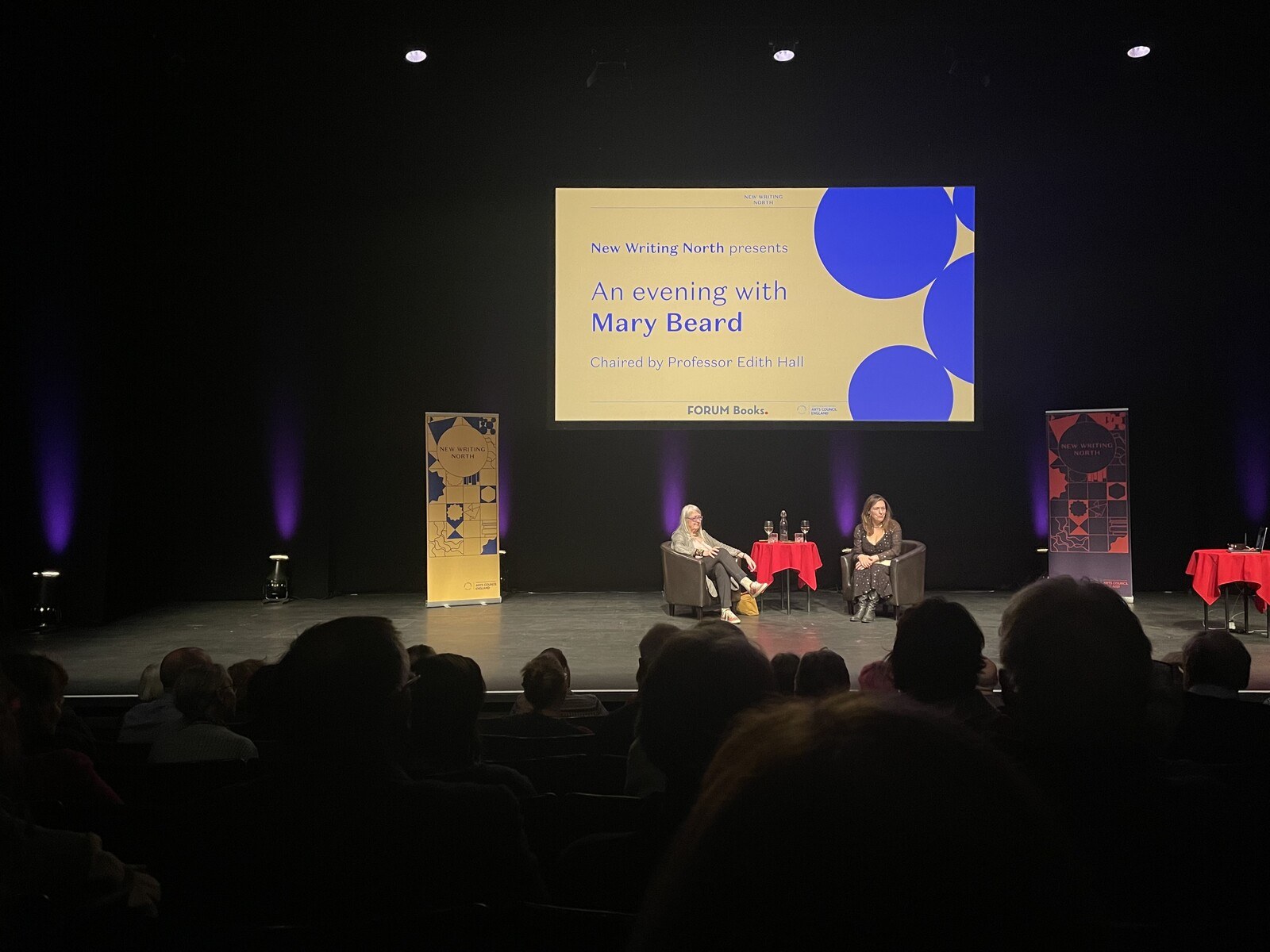 Mary Beard and her interviewer on stage in front of a sign reading "An evening with Mary Beard"