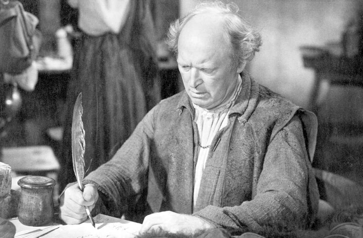 A man with light hair and a worried expression sits at a desk, holding a feather quill and writing on parchment. In the background, a woman in a dress is partially visible. From Paracelsus (1943).
