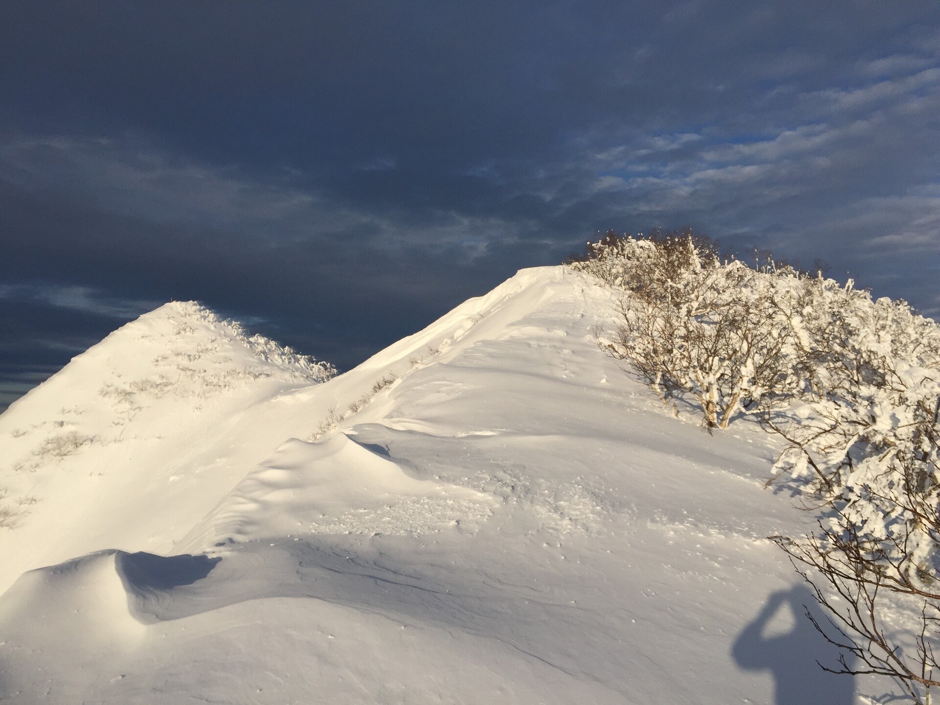 A snowy ridgeline with the sun upon it; dark clouds in the sky beyond
