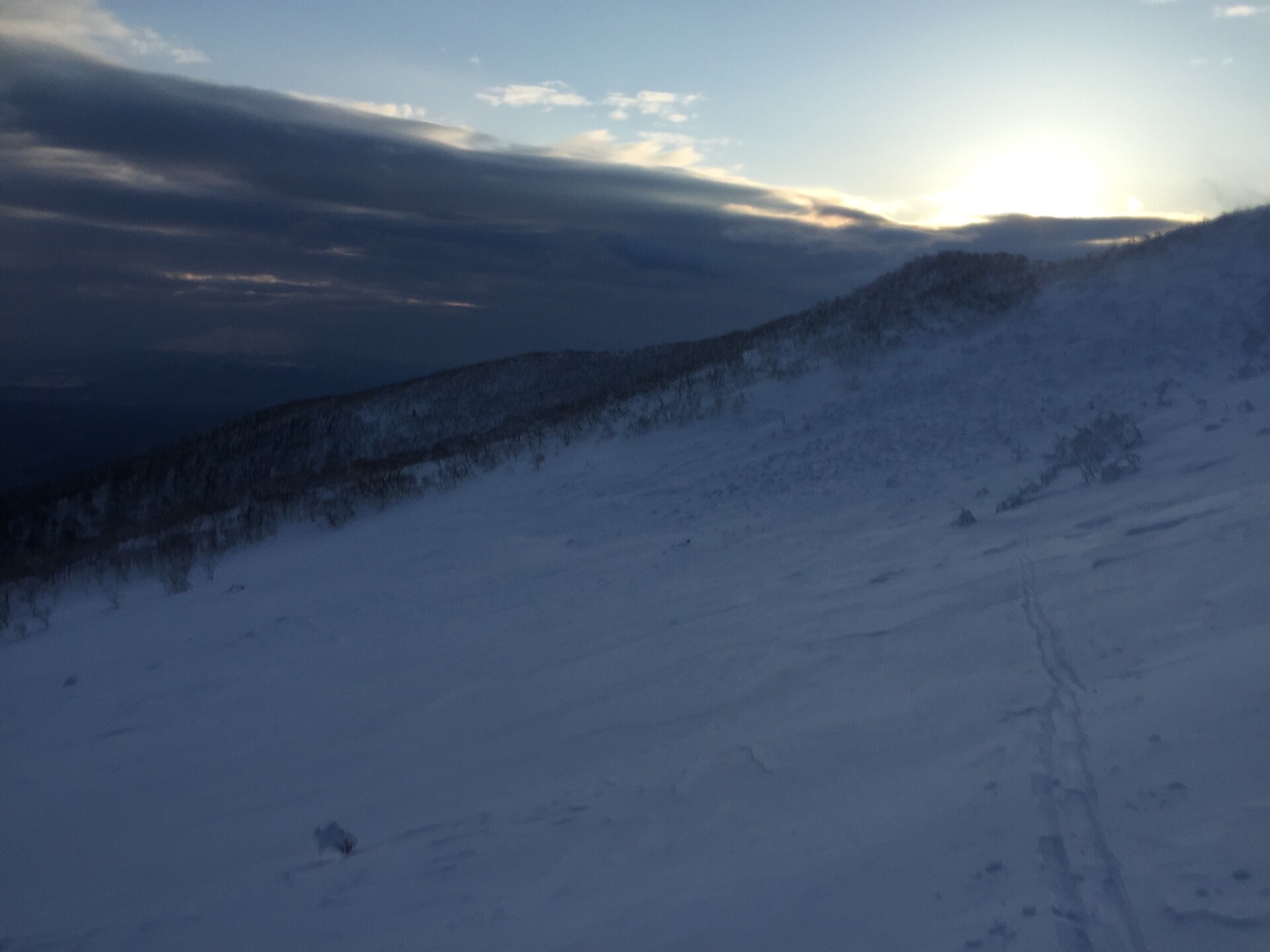 Looking down a windblown, treeless slope, covered with snow, dark clouds on the left and a sunrise on the right