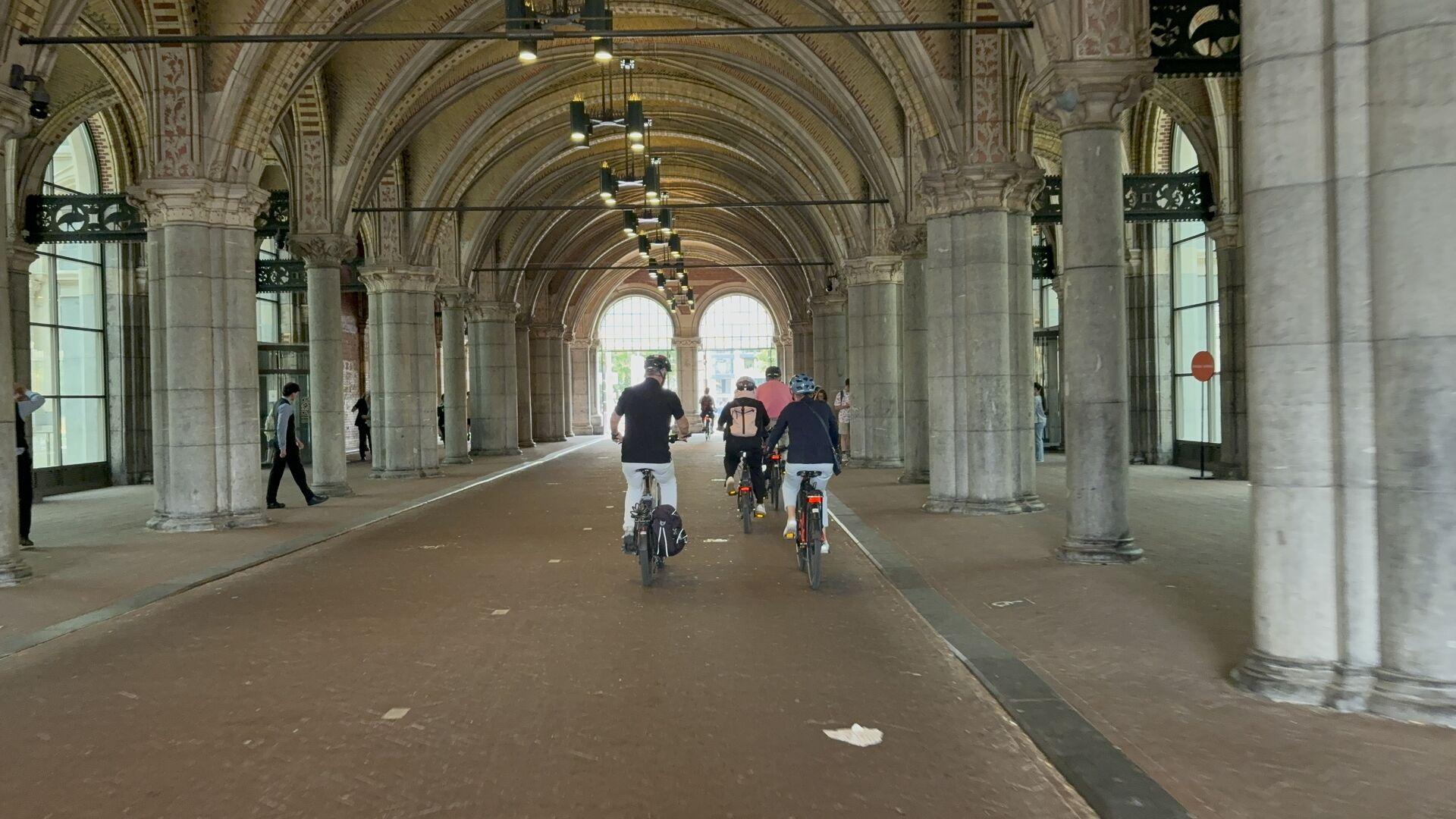 People ride bicycles under an arched stone arcade with ornate columns and hanging lights, near large windows and doors on both sides.