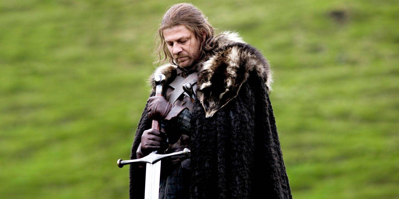A somber man dressed in medieval armor and a dark, fur-lined cloak stands in front of a blurred green landscape. He holds a sword in front of him, looking down thoughtfully. His long hair falls around his face, and a serious expression is evident as he reflects on something significant. It's Ned Stark from Game of Thrones
