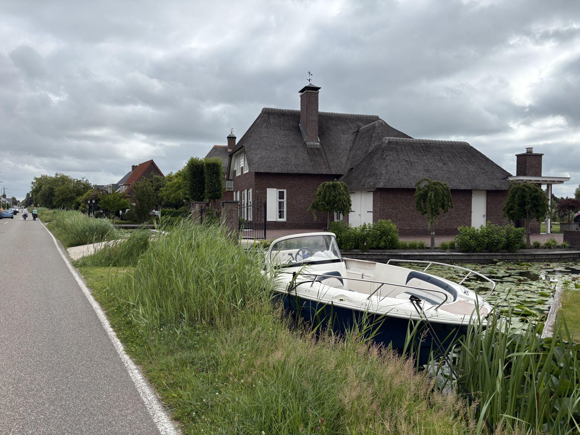 Street view with a boat docked in a narrow canal in front of a brick house with a thatched roof. Overcast sky, grass and lily pads line the canal, with cyclists on the road.