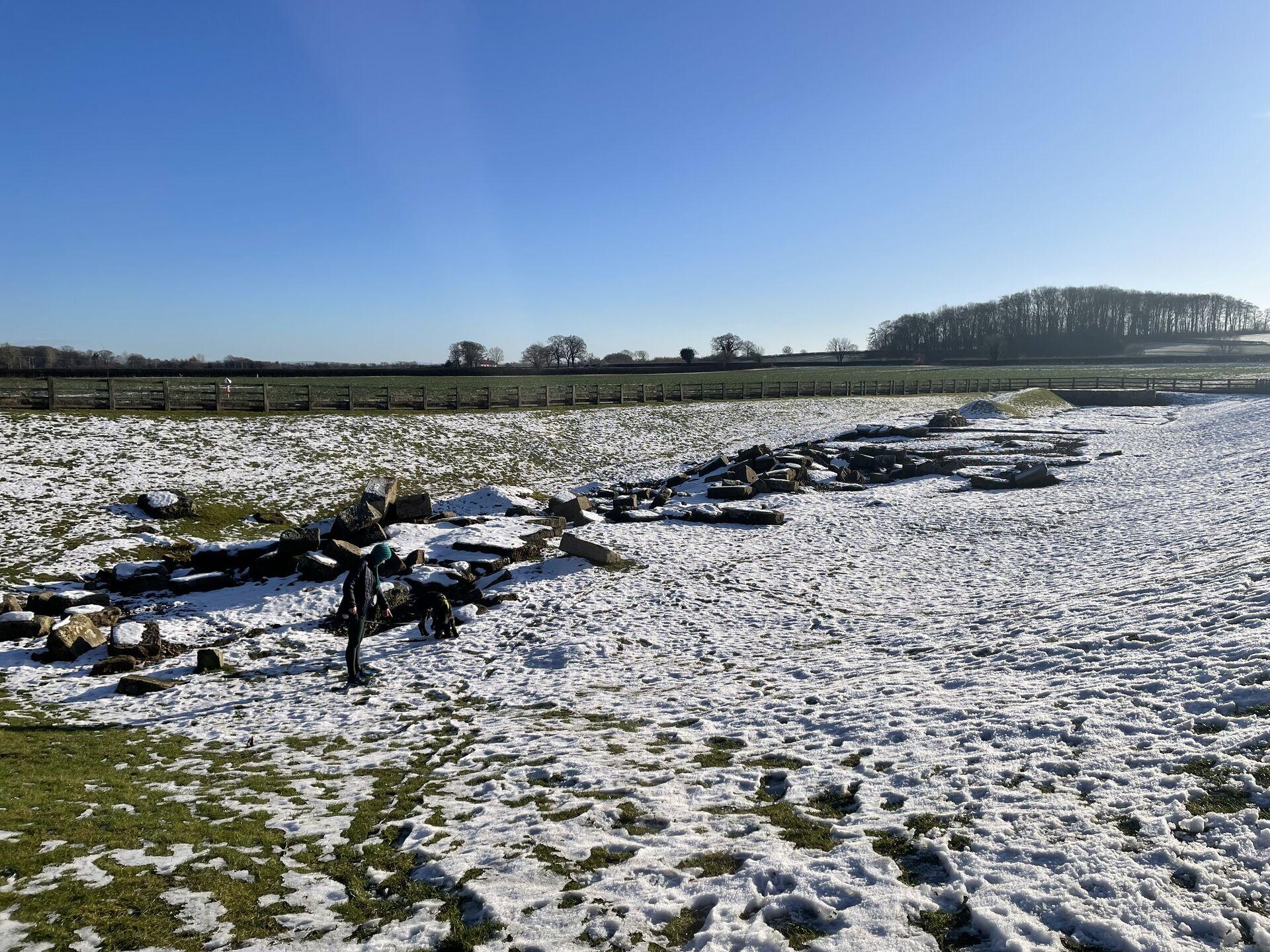 A clear day at the Piercebridge Roman Bridge, set in a depression in a grassy field, covered in snow