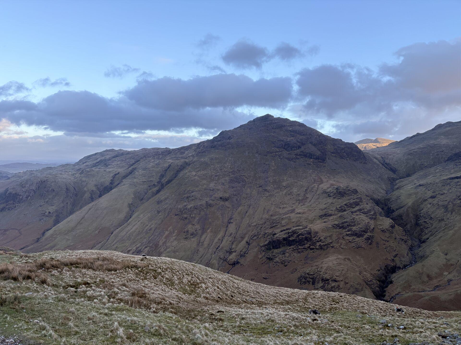 A high, pyramidal mountain seen from a grassy hillside in the foreground; crags at its summit and little ravines on its grassy flanks below.
