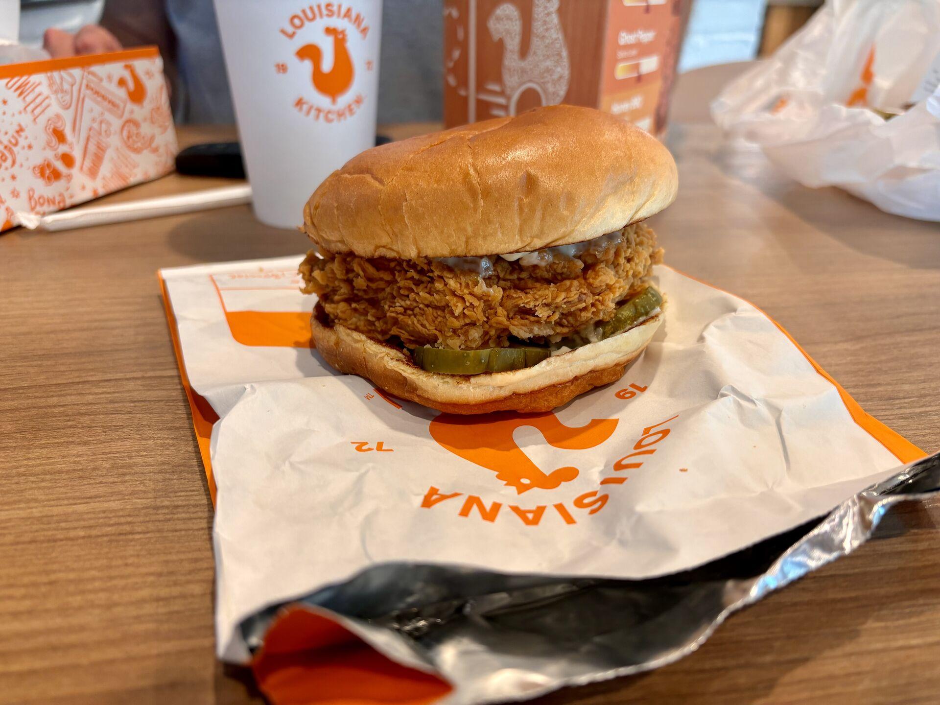 A chicken sandwich literally bursting with pickles on a paper bag, with various other Popeyes memorabilia in the background.