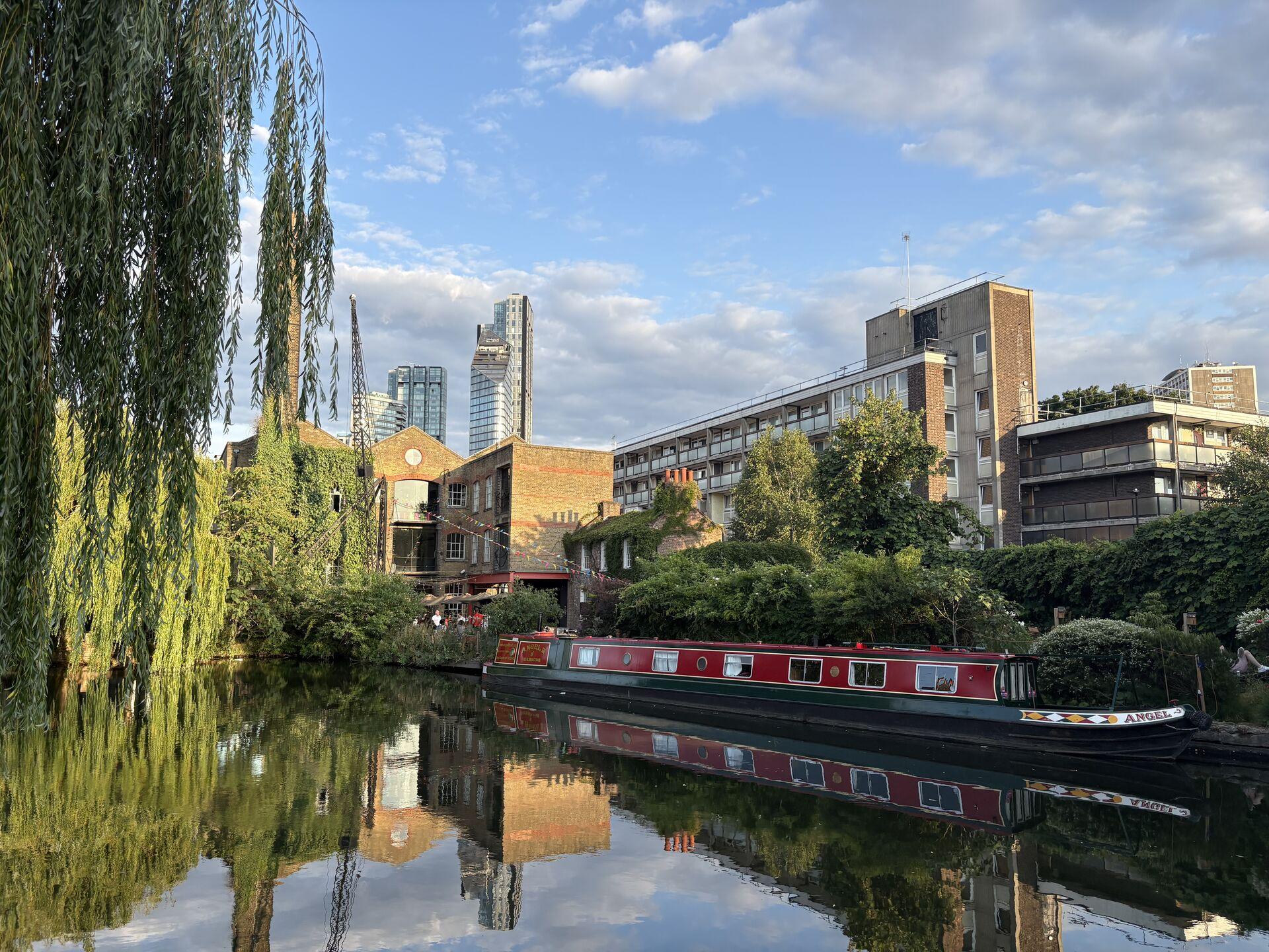 Canal boat named "Angel" moored along a canal with brick buildings, modern skyscrapers in the background, and weeping willow trees hanging over the water reflecting the scene under a partly cloudy blue sky.