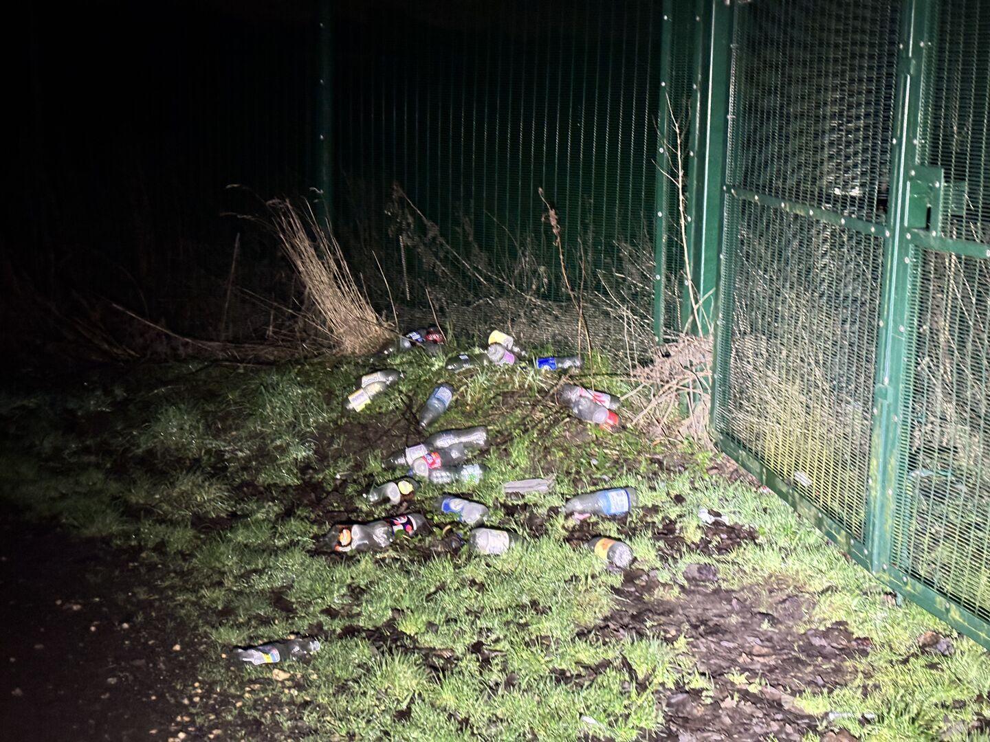 A pile of discarded plastic bottles sitting in the grass next to a fence, illuminated by a flashlight beam