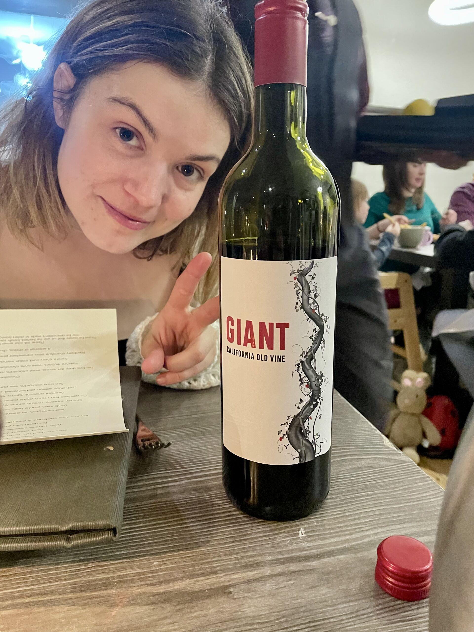 A young woman with shoulder-length hair smiles and makes a peace sign next to a bottle of red wine labeled "GIANT California Old Vine." A menu is partially visible on the table.