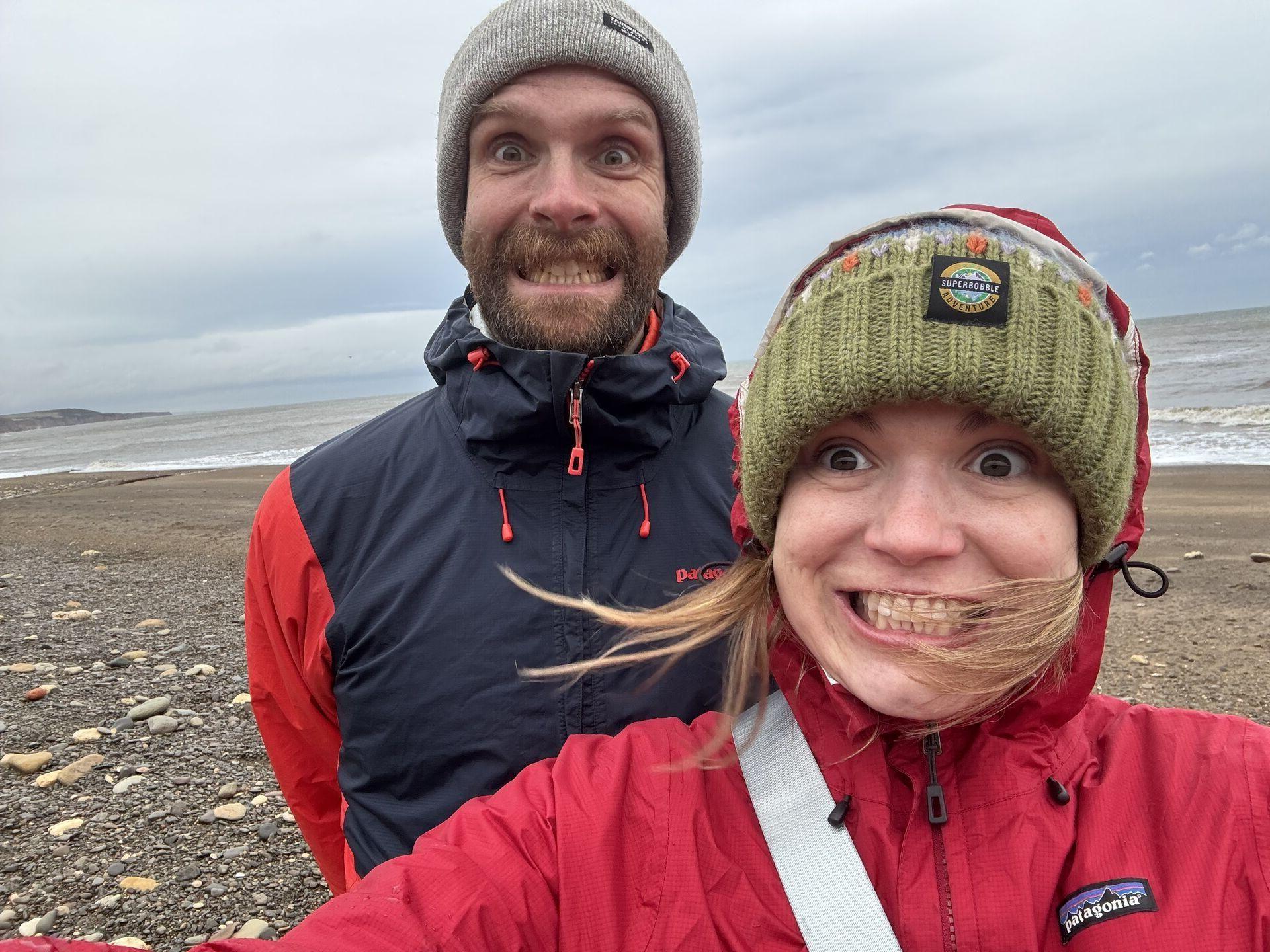 A man and a woman stand on a beach, both making exaggerated, playful facial expressions. They are wearing warm jackets and hats, with windblown hair. The ocean and cloudy sky are visible in the background.