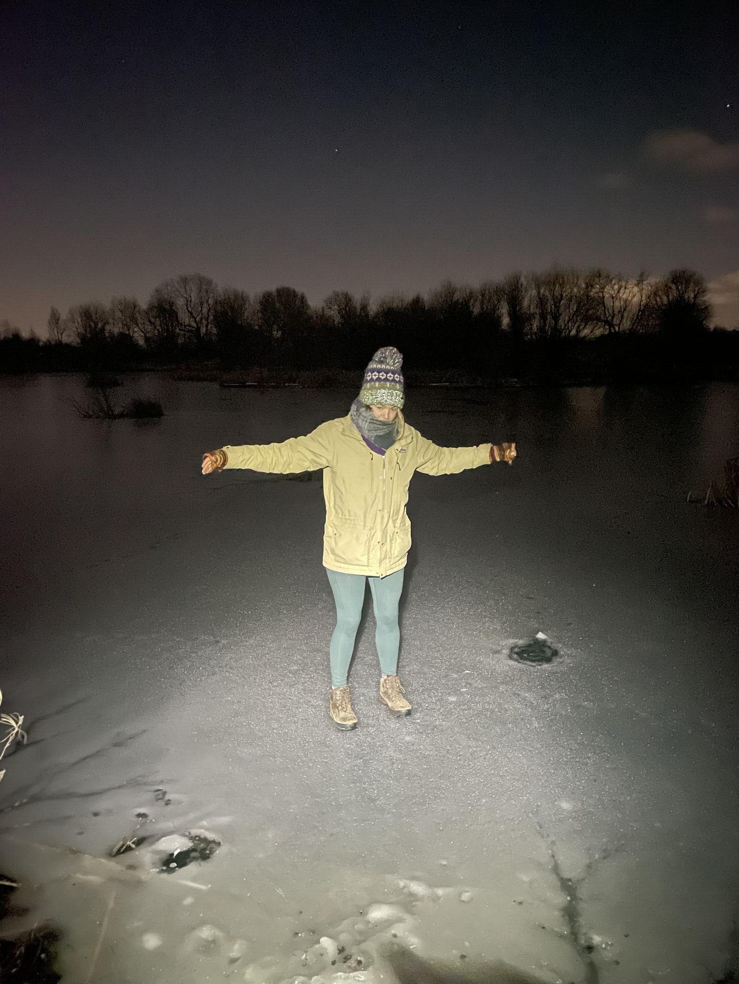 A woman bundled up in warm clothes standing on a frozen-over lake in the dark. Her arms are splayed for some reason