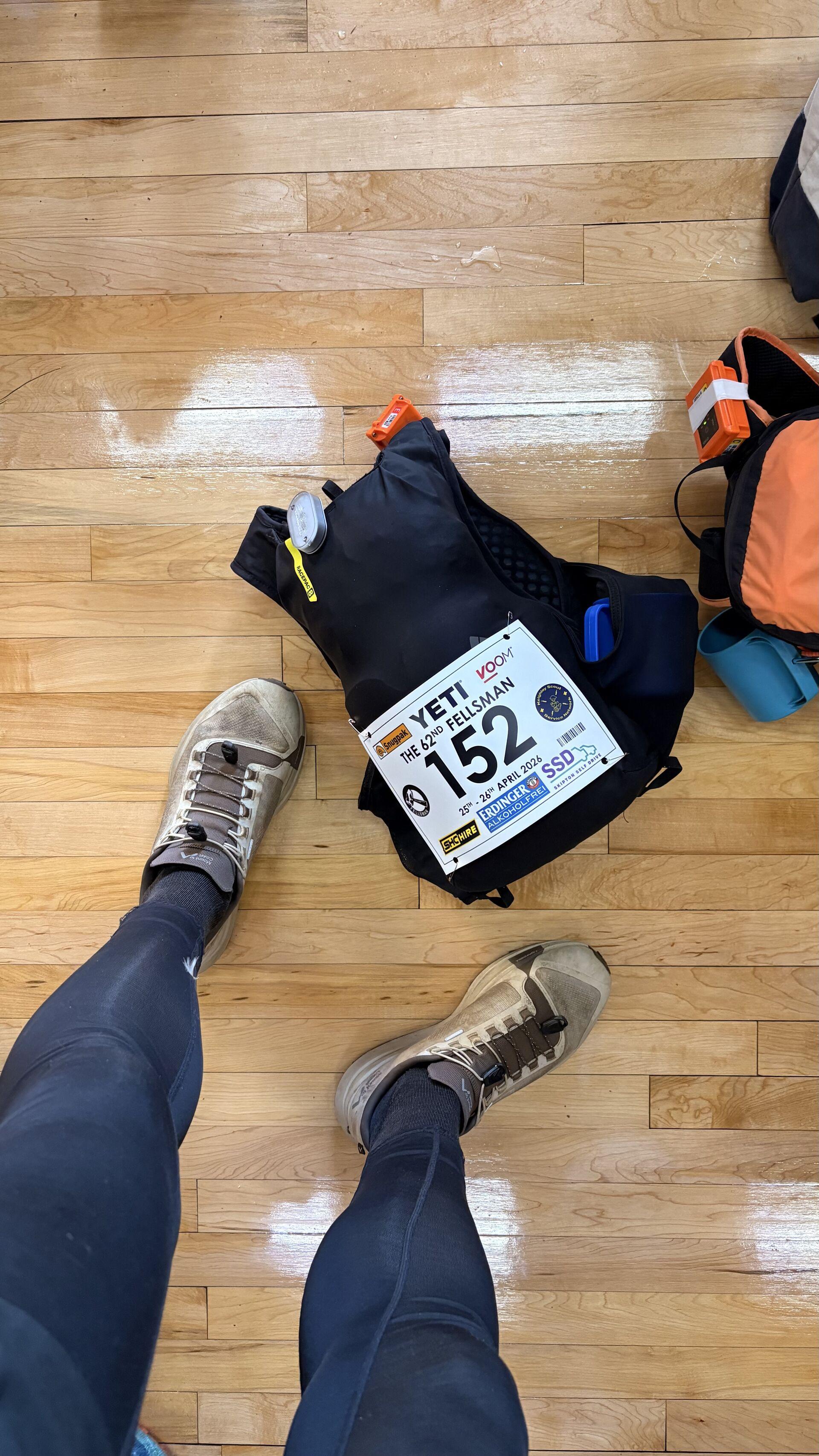 A point of view shot of a pair of legs standing over a running backpack with the number 152 pinned on, on a wooden floor
