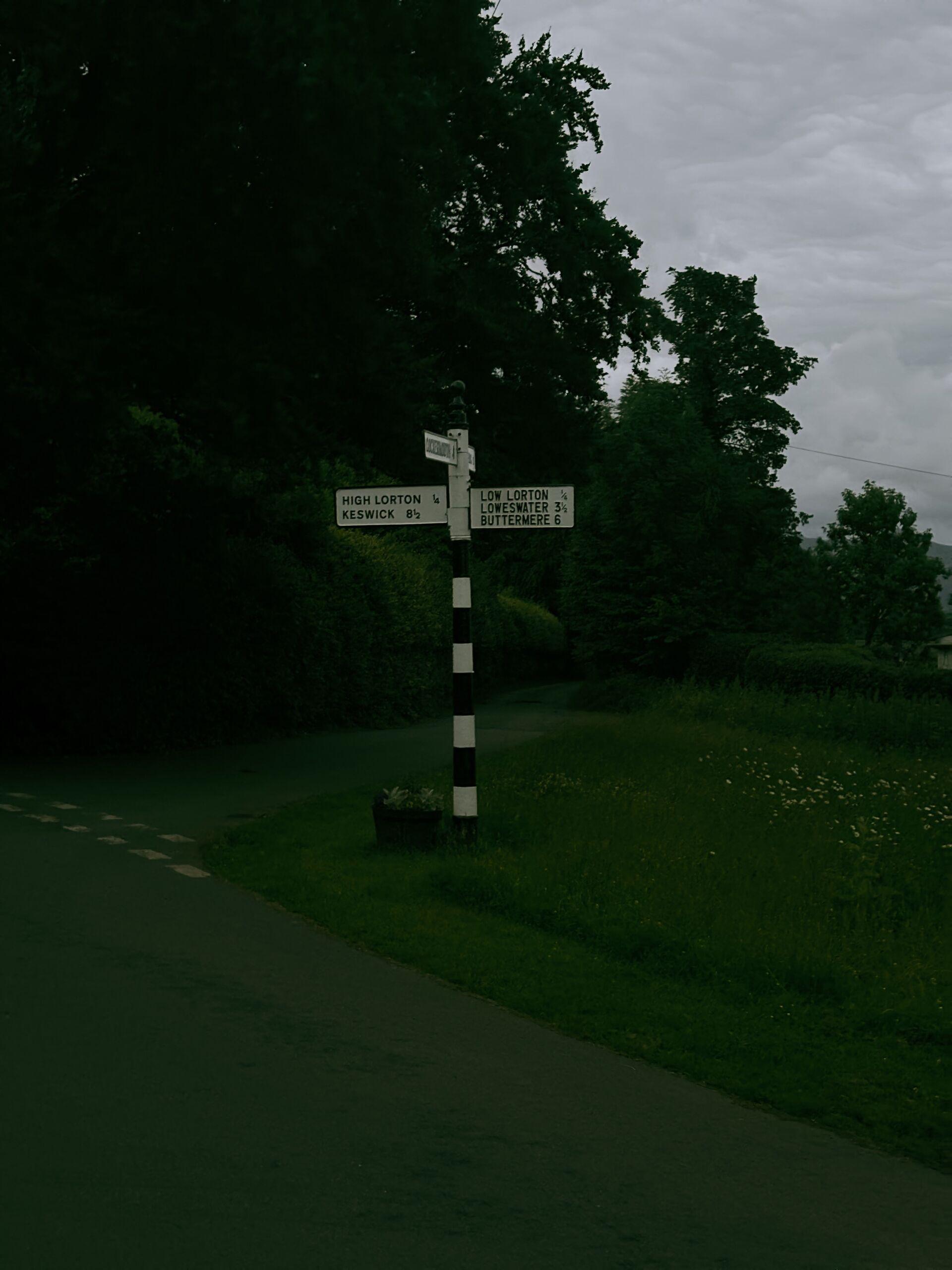 A black-and-white striped road signpost at a rural junction pointing to High Lorton, Keswick, Low Lorton, Loweswater, and Buttermere, surrounded by trees and hedgerows.