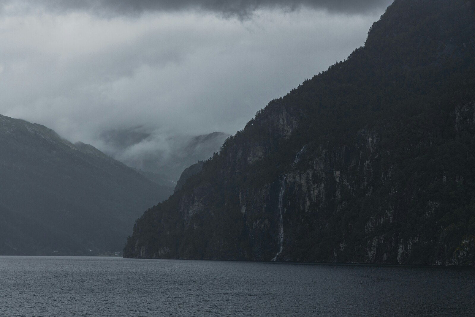 A dark sky over a rocky cliff descending into the grey waters of a fjord