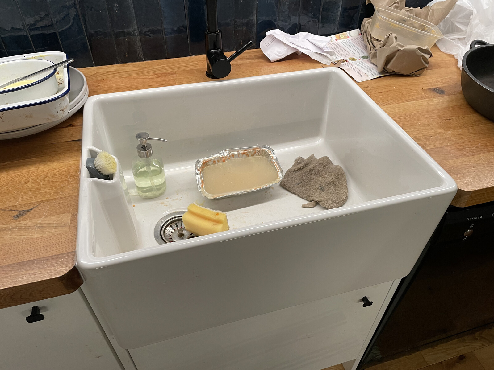 An apron-fronted sink in a wood countertop with a bunch of rubbish & dirty dishes lying around it