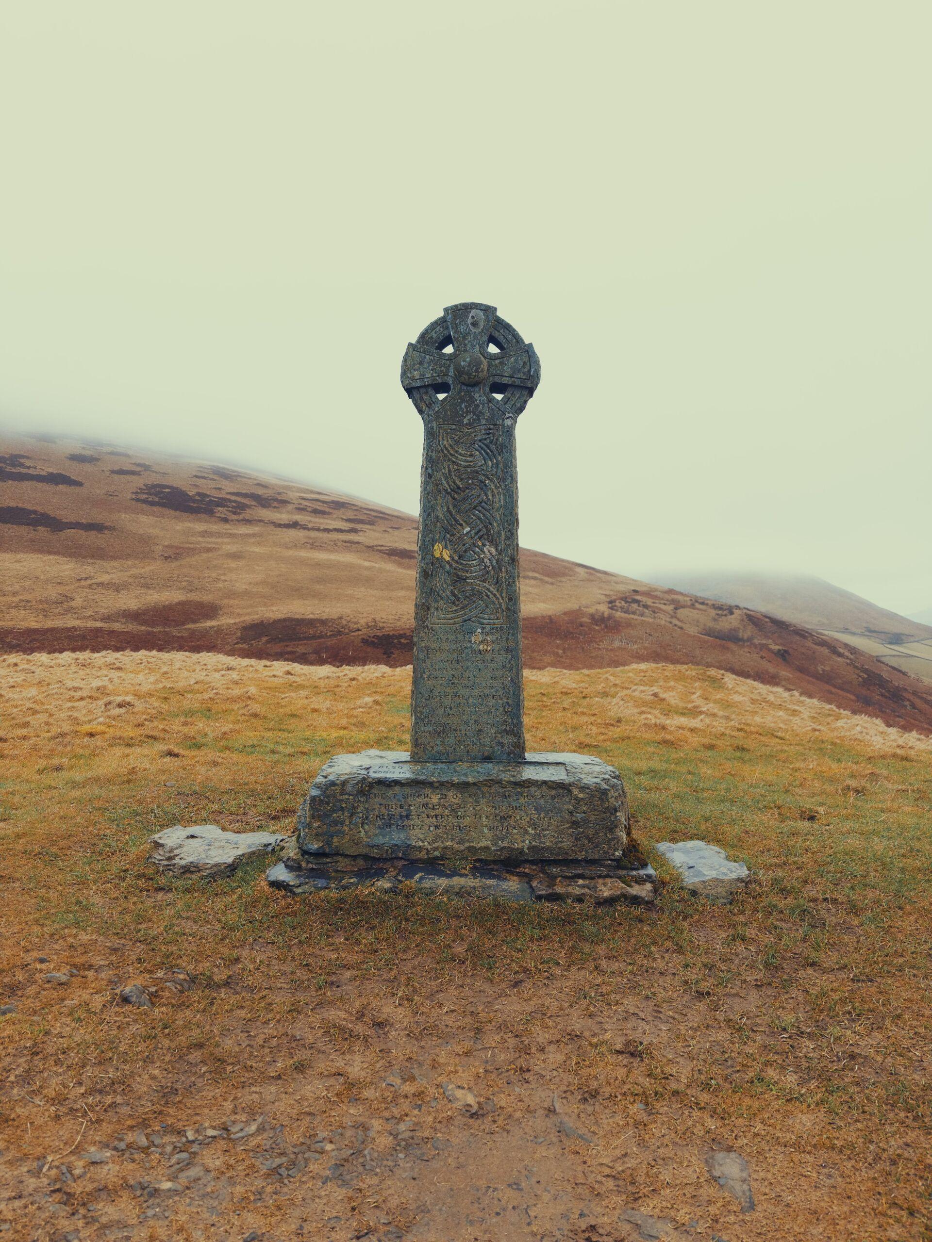 A celtic cross on a stone plinth memorialising two shepherds, on a grassy hillside with a moor rising in the background beyond