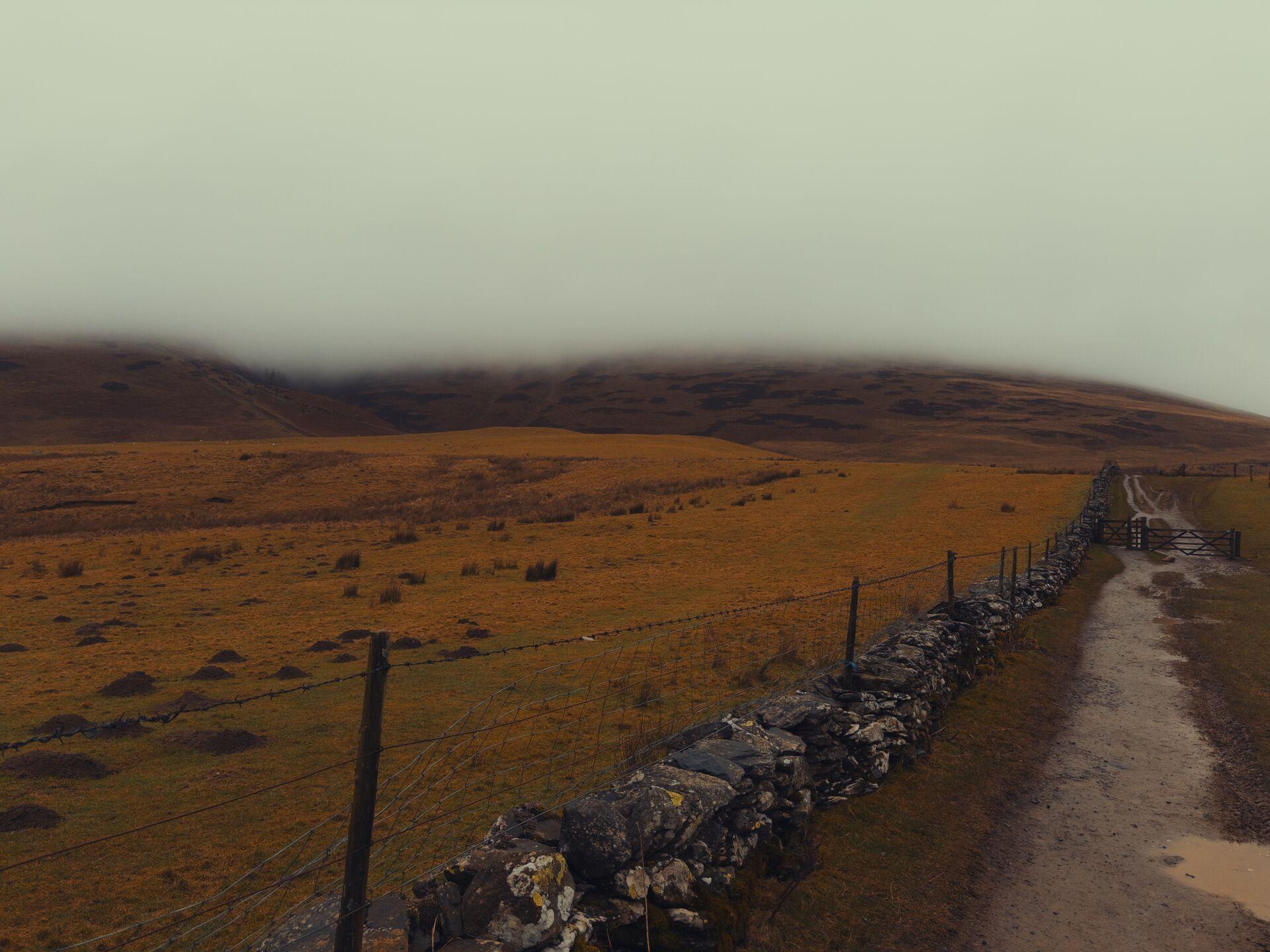 A track follows a low wall and wire fence into the distance, with the bulk of a mountain rising on the left into the clouds.