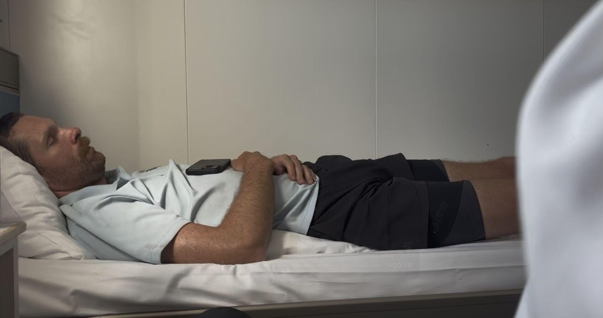 Man with beard lying on a bed with eyes closed, wearing a light-colored shirt and dark shorts, with a phone resting on his chest on the berth of a cabin on a ferry.
