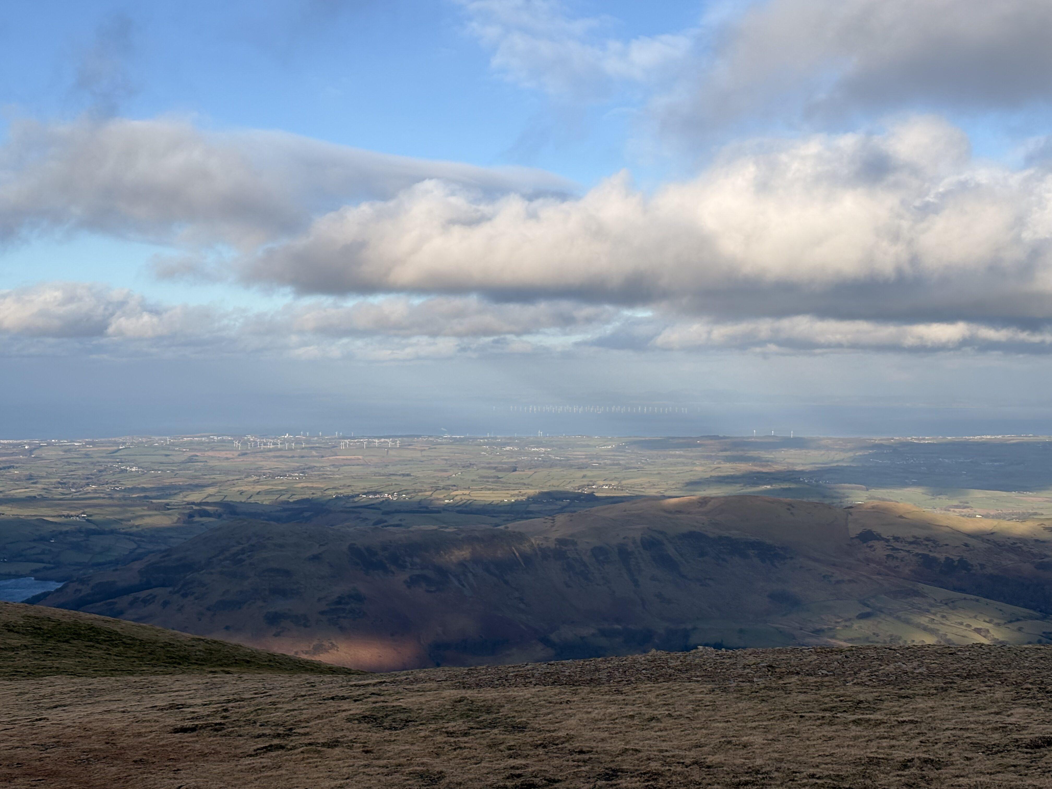 A view of fells in the foreground and low green fields rolling off towards the sea, under a patchy sky. There's a windfarm off the coast in the far distance