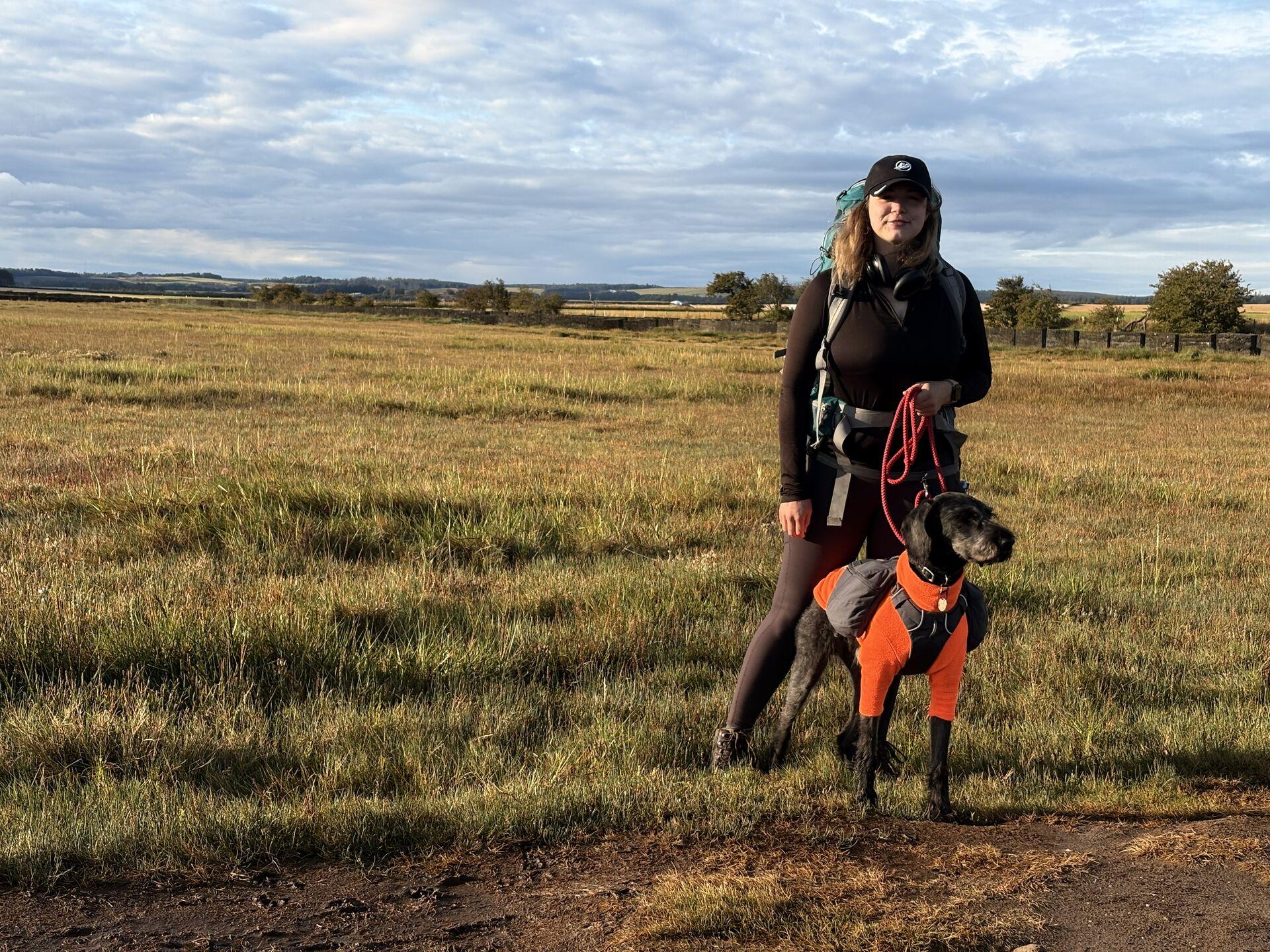A woman and a dog wearing an orange fleece standing in a field of tussock grass in the gold light of a sunrise