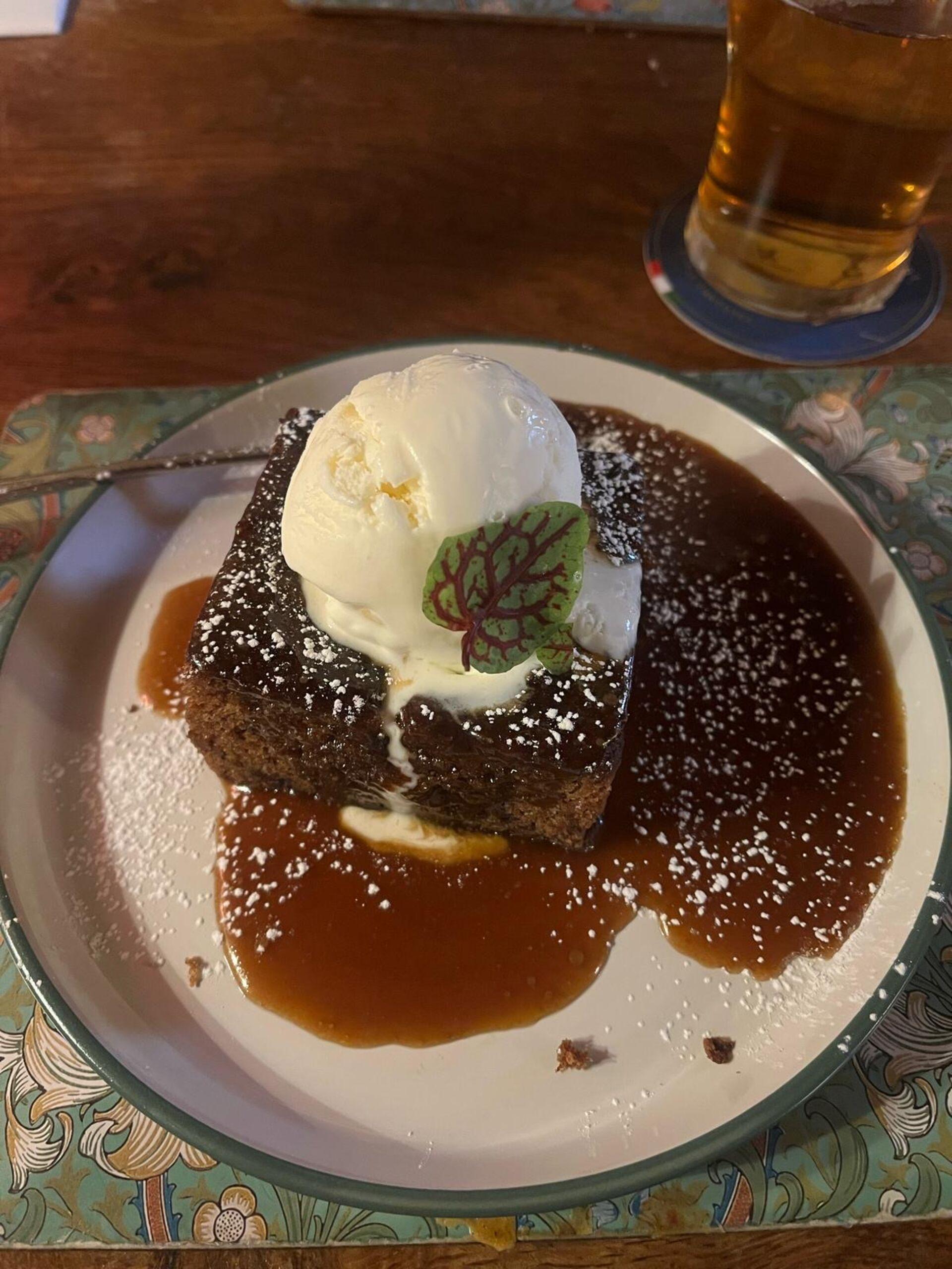 A sticky toffee pudding, topped with ice cream and heaped with caramel sauce, with a beer in the corner of the frame