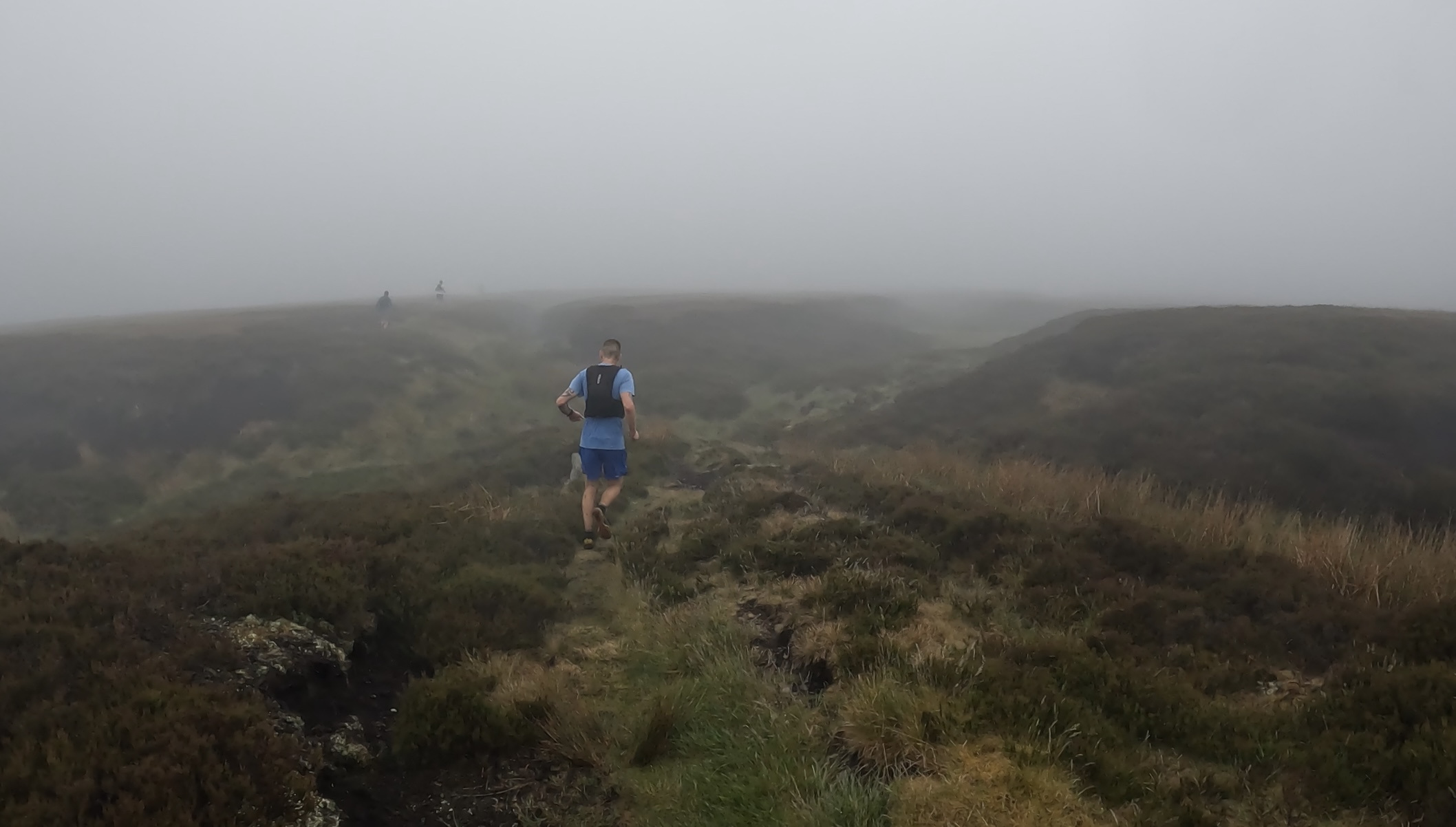 Fell runners cross an open moor, following a track amid heather, and all socked in with cloud
