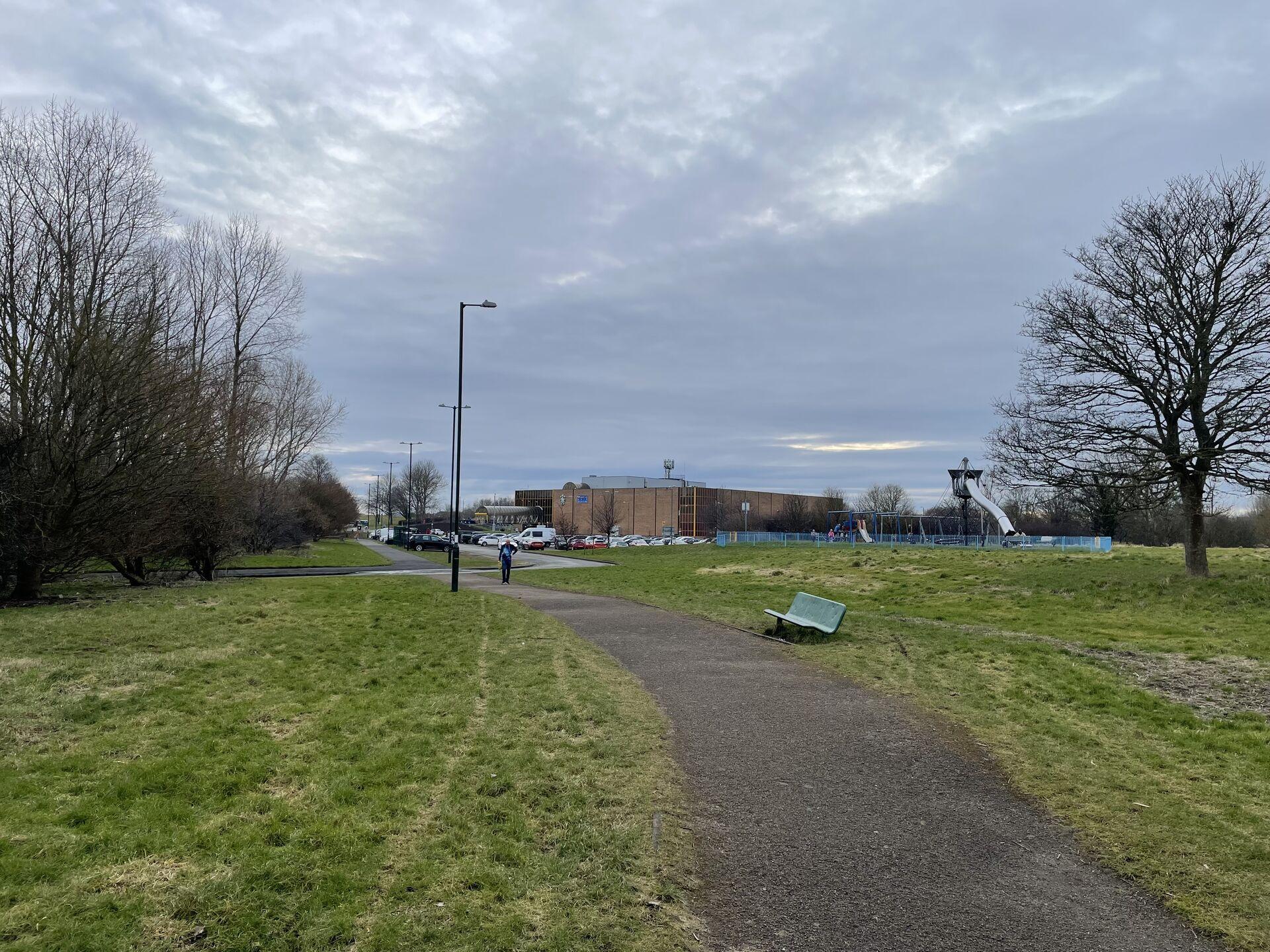 A squat brick building, maybe from the 1970s, stands by a parking lot and a tarmac path leading through grassy park fields