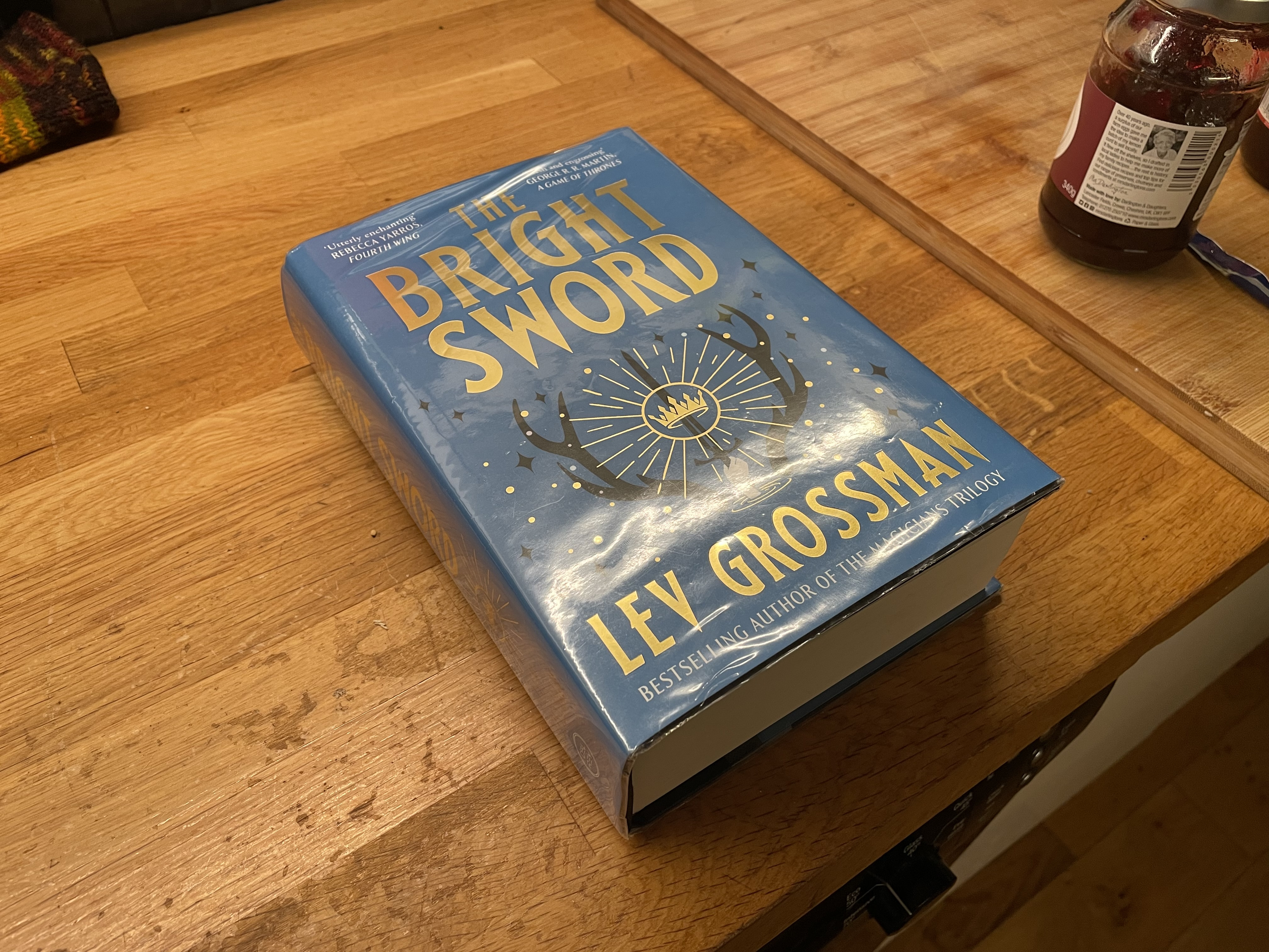 The Bright Sword, by Lev Grossman, sitting on my wood countertop next to a jar of raspberry jam.