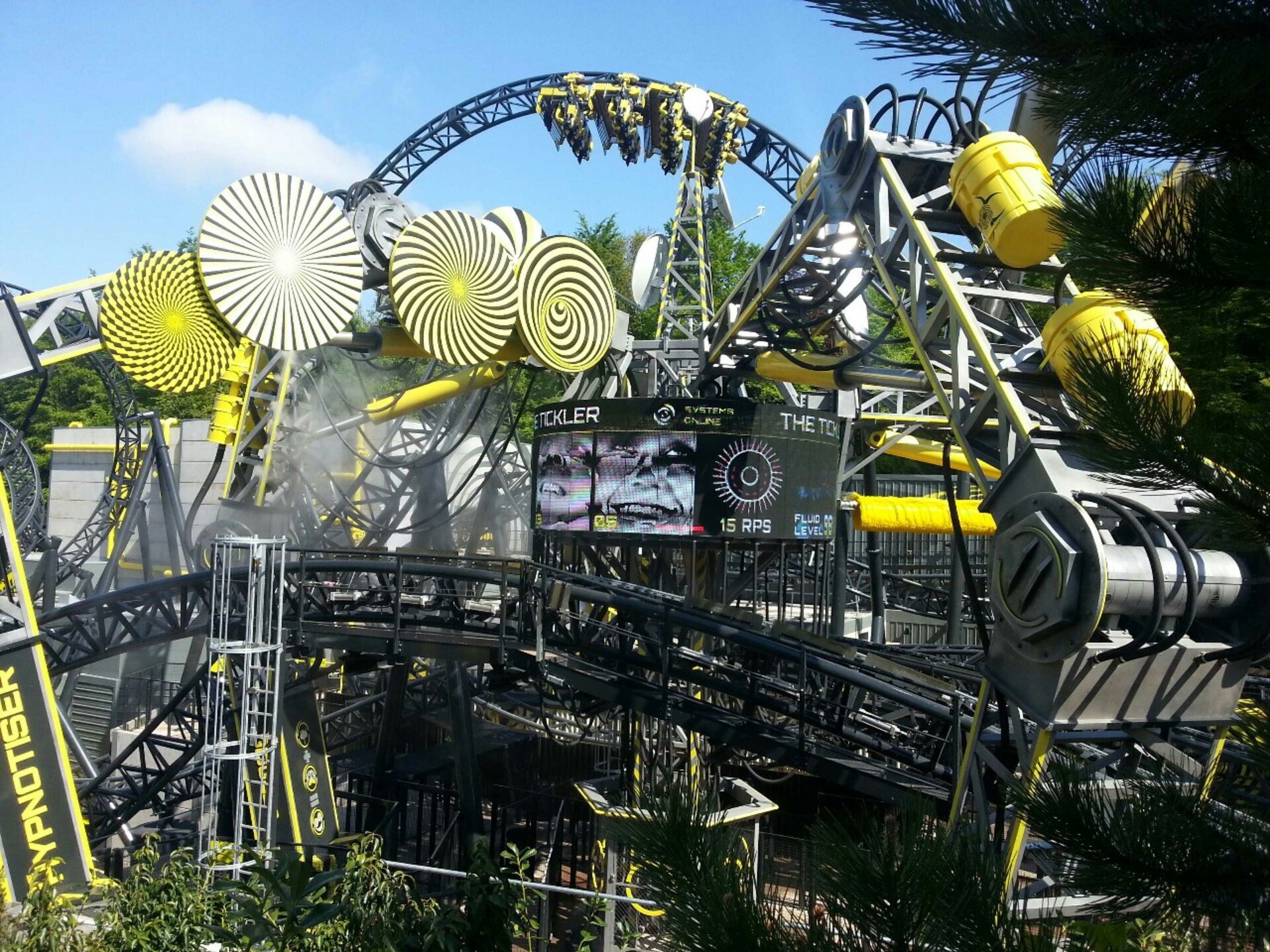 A roller coaster in grey and yellow, with tons of bright spirals on panels and some big screens in the middle showing people smiling