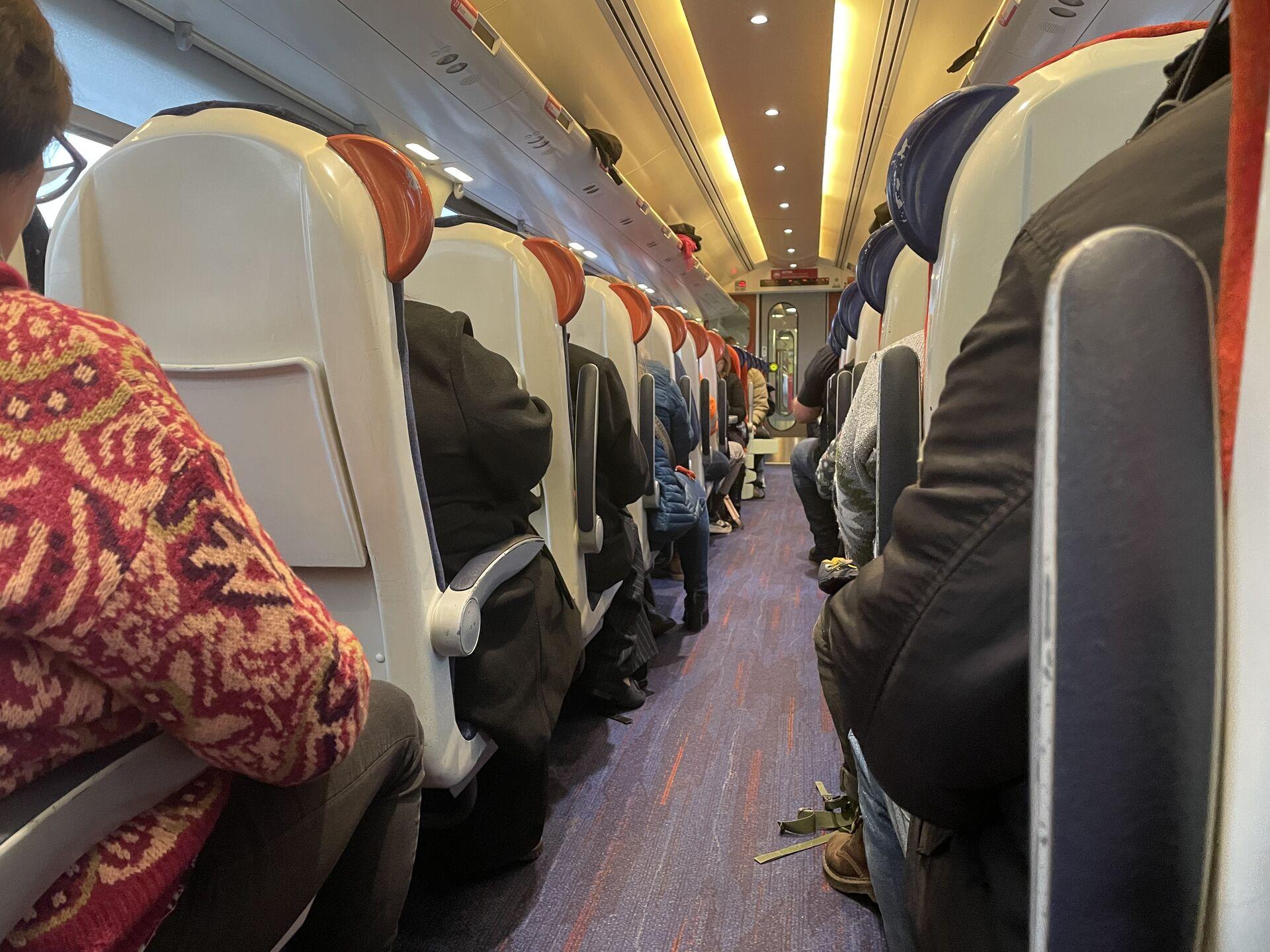 Interior view of a train showing rows of seats and passengers sitting with their backs to the camera. A person in a patterned sweater is visible in the foreground. Soft lighting illuminates the aisle.