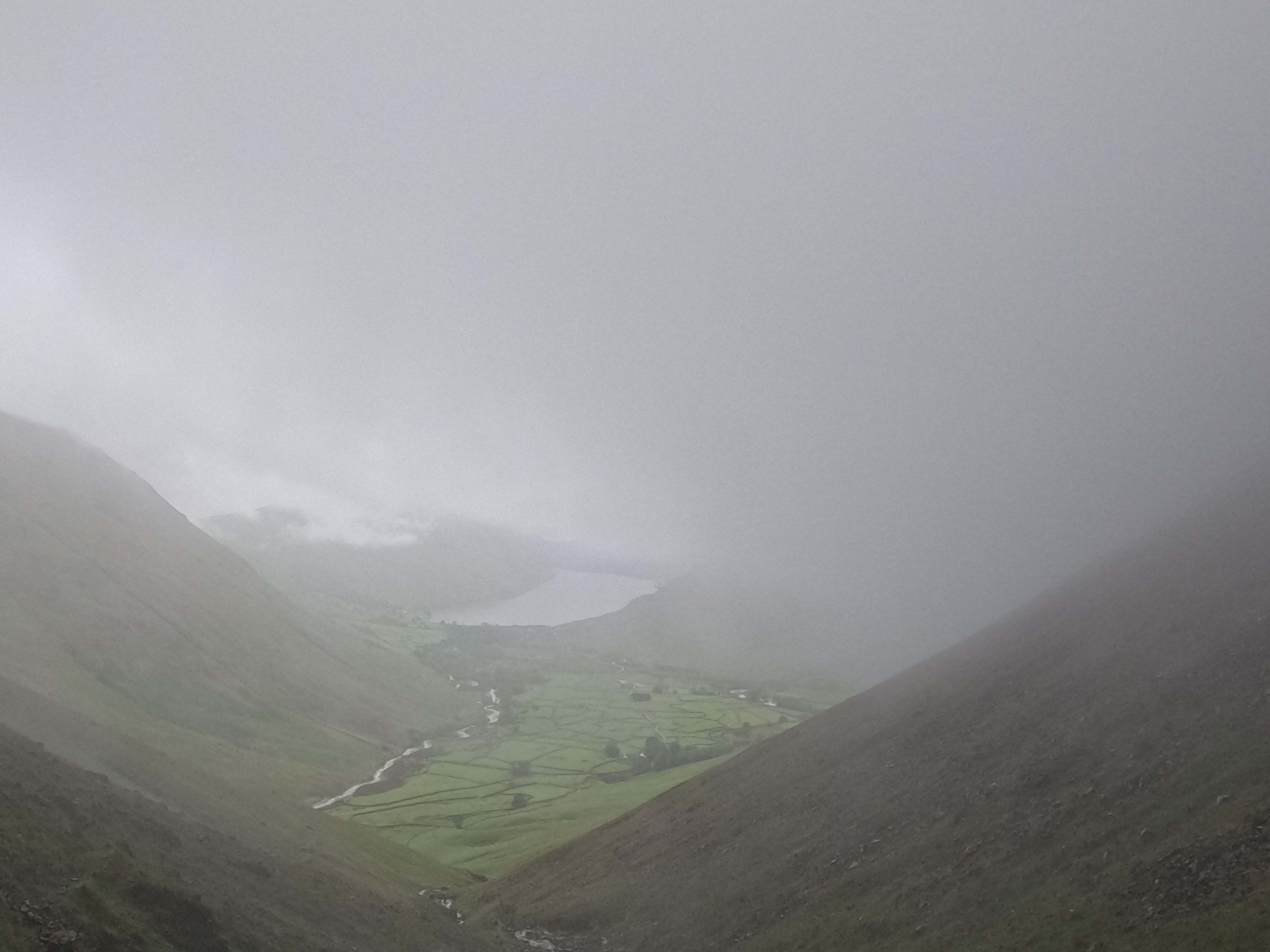 Looking through clouds at a green valley below, with dark valley walls rising on either side, and some small stone walls visible in the distance through the mist