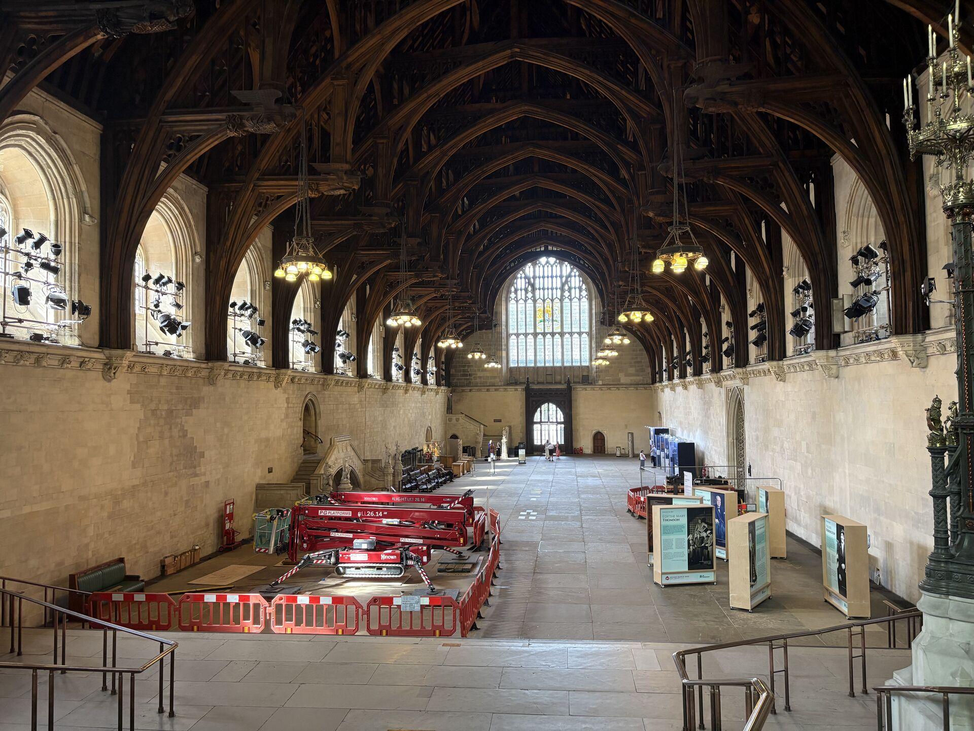 Interior of a large historic hall with wooden vaulted ceiling, stone walls, large arched windows, red construction barriers and equipment, and several informational display boards on the right side.