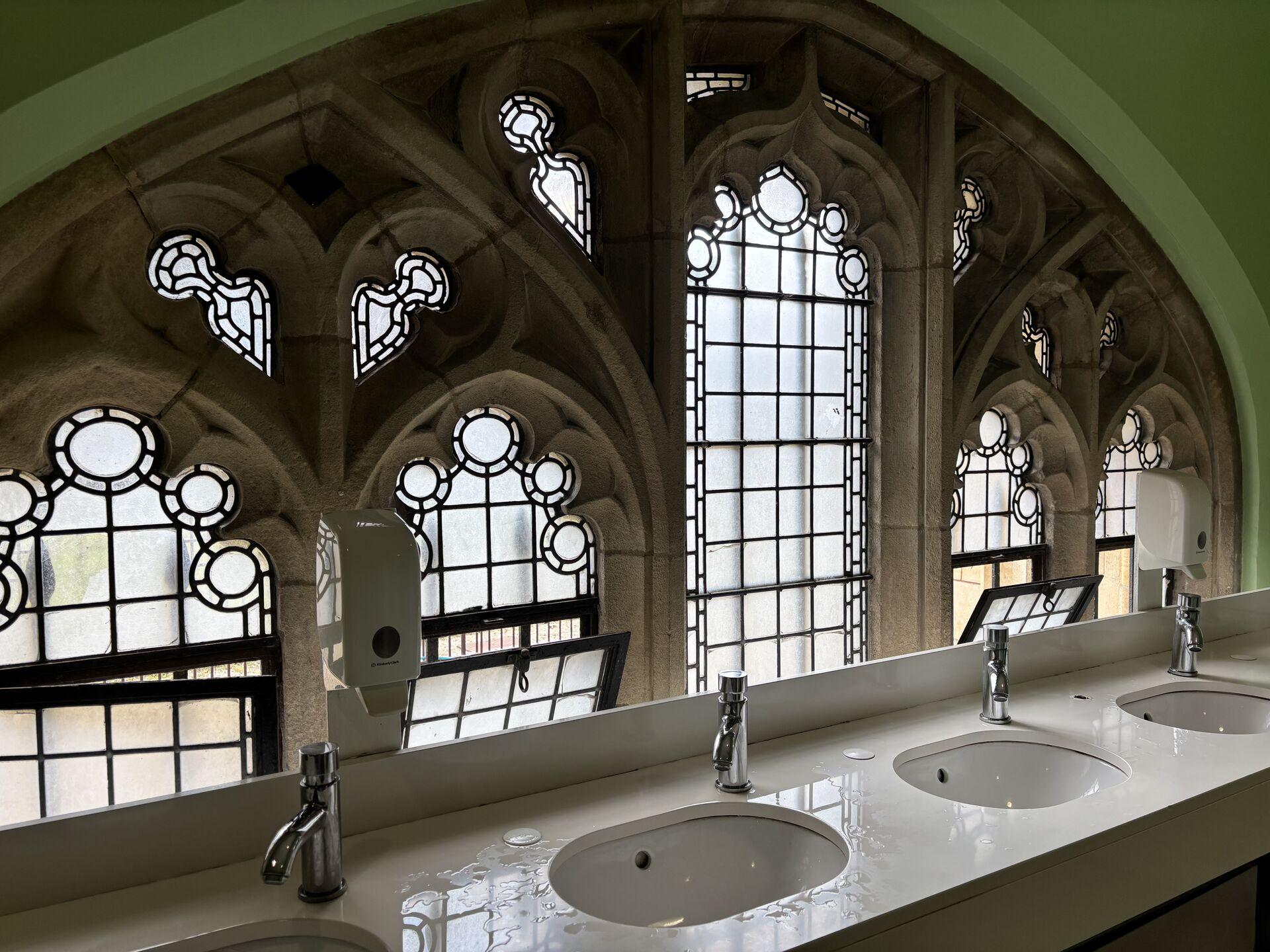 Modern white sinks with metal faucets under ornate gothic-style stained glass windows with geometric patterns in stone frames, inside a restroom with green walls.